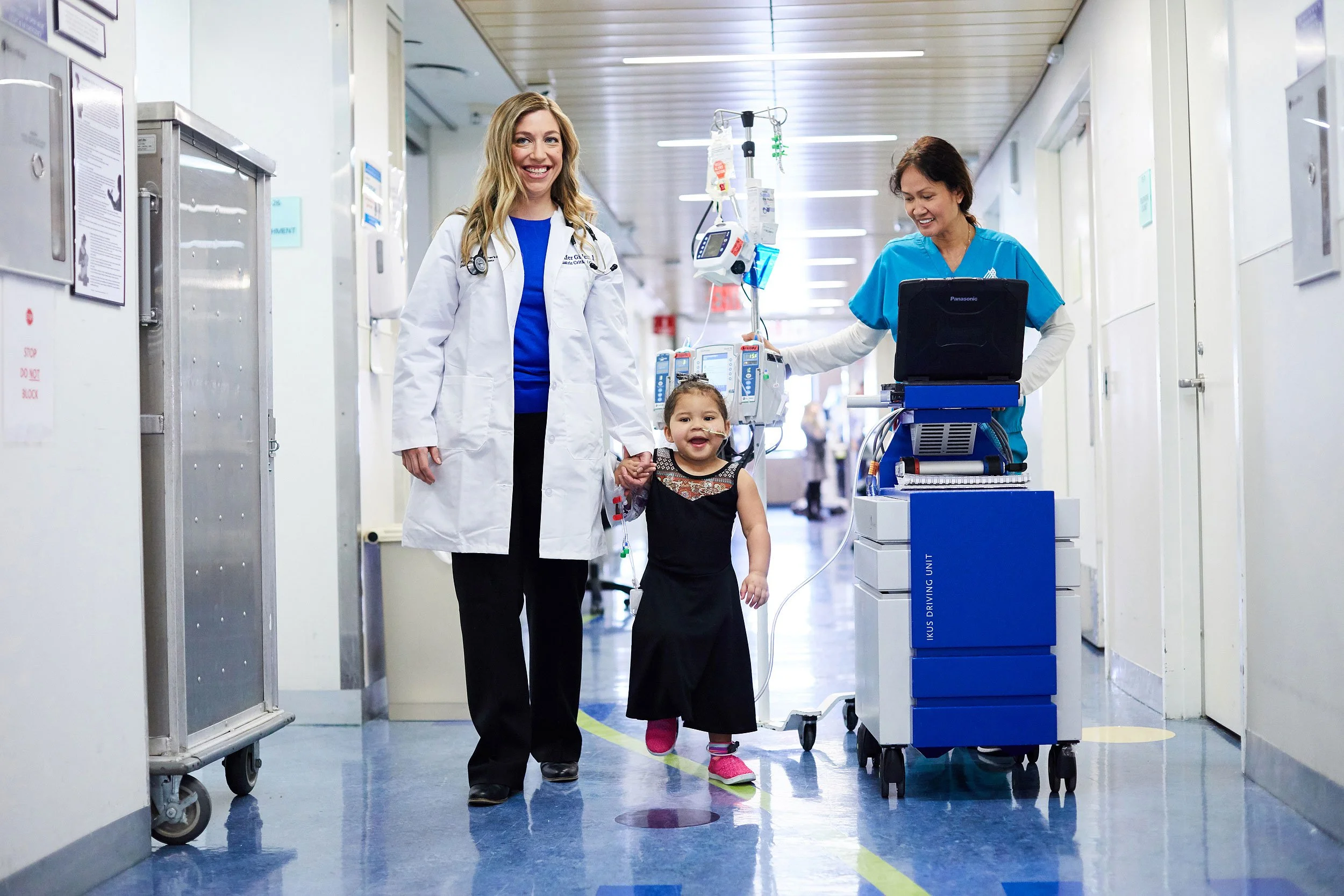 Doctor, nurse and young child with IV walking together down a bright hospital hallway.
