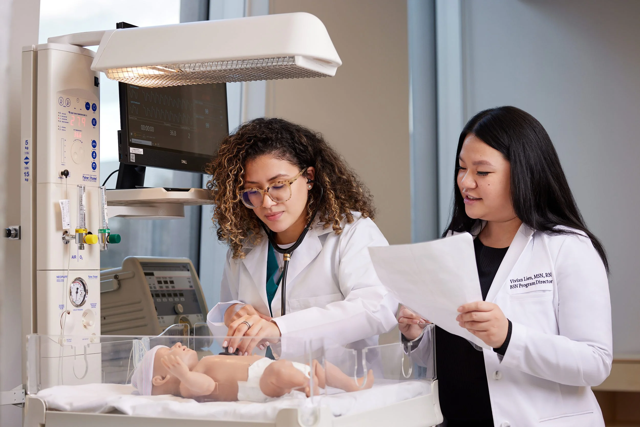 Female in labcoat demonstrating neonatal equipment to a colleague in a training facility.