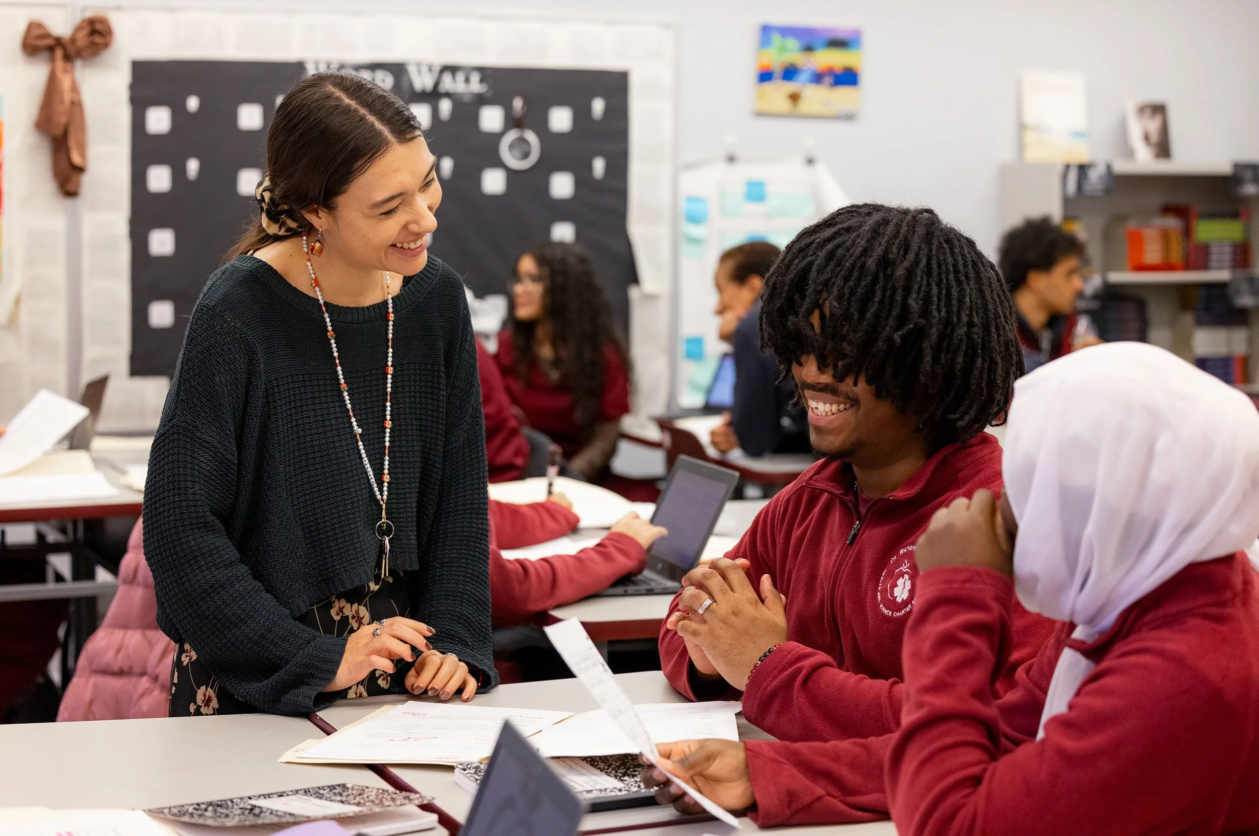 Two students interacting with teacher in classroom setting, smiling. 