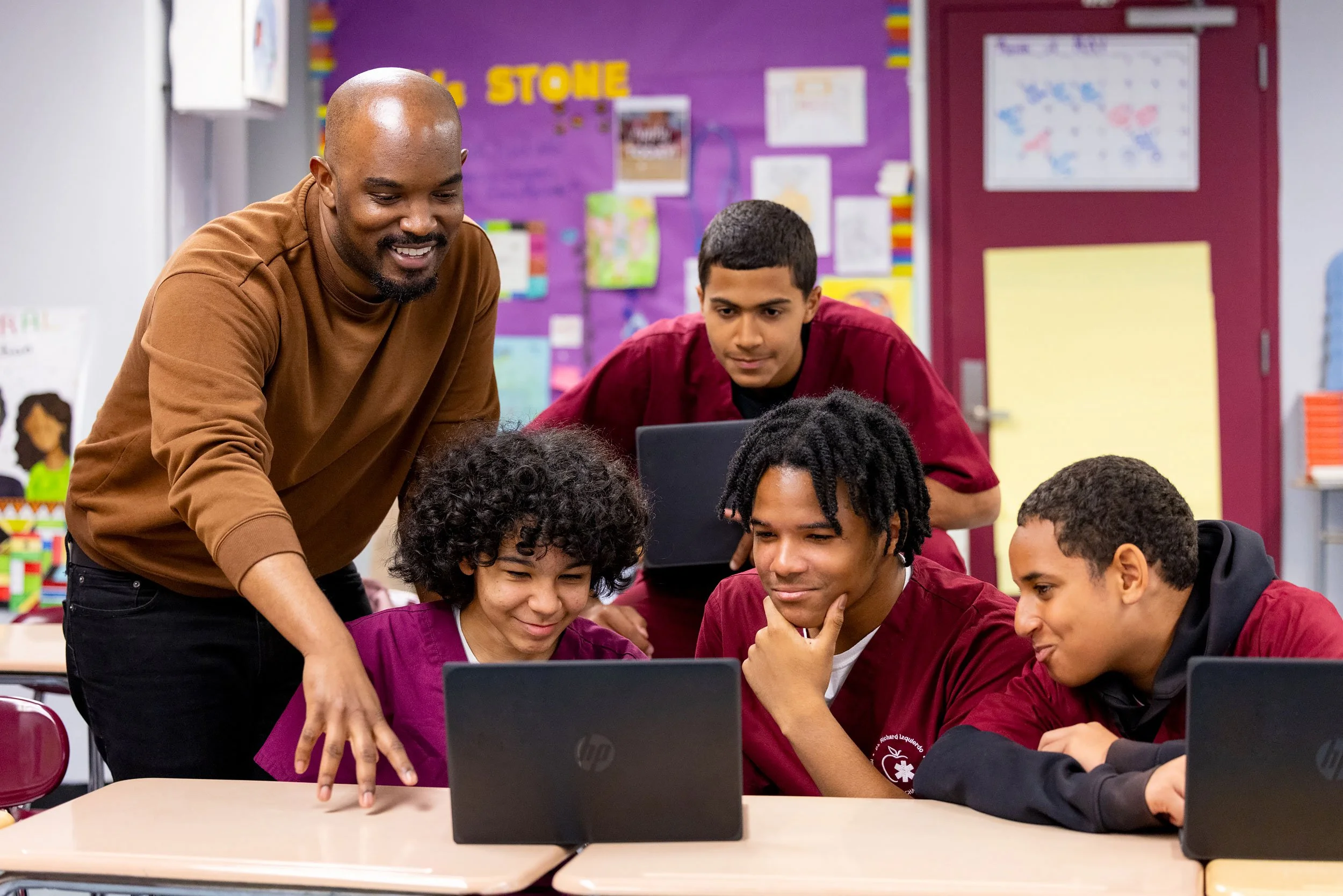 Teacher assisting 4 students working together at table with laptops.