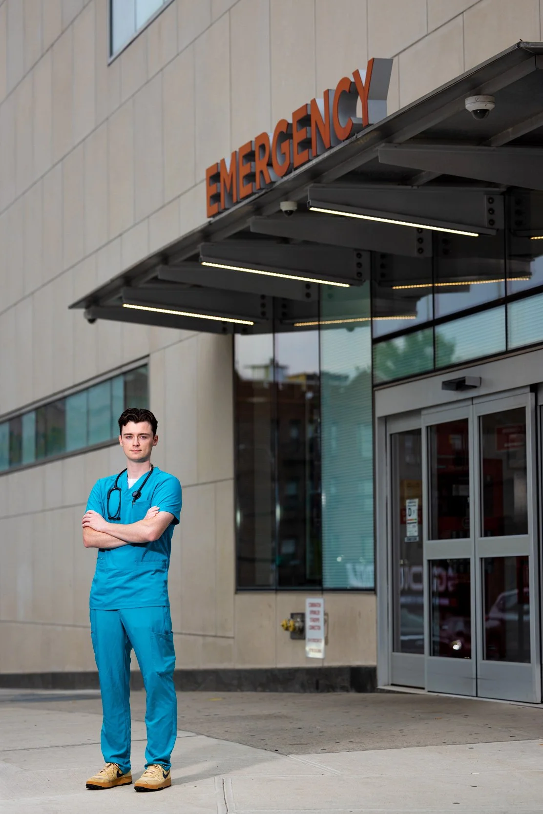 Male healthcare worker in teal scrubs standing outside a hospital emergency department entrance.