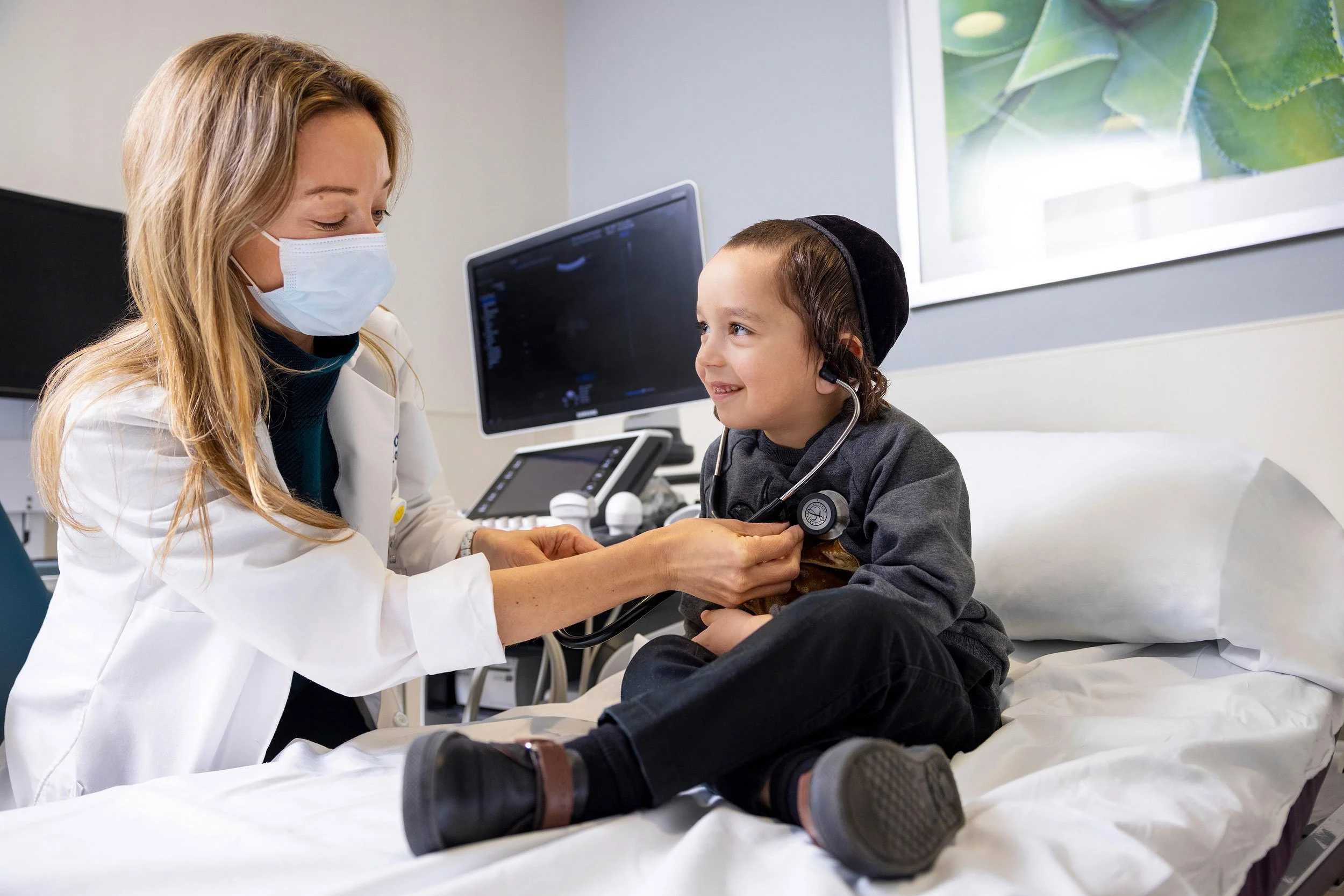 Female pediatrician with surgical mask listening to a young boy’s heartbeat with a stethoscope.