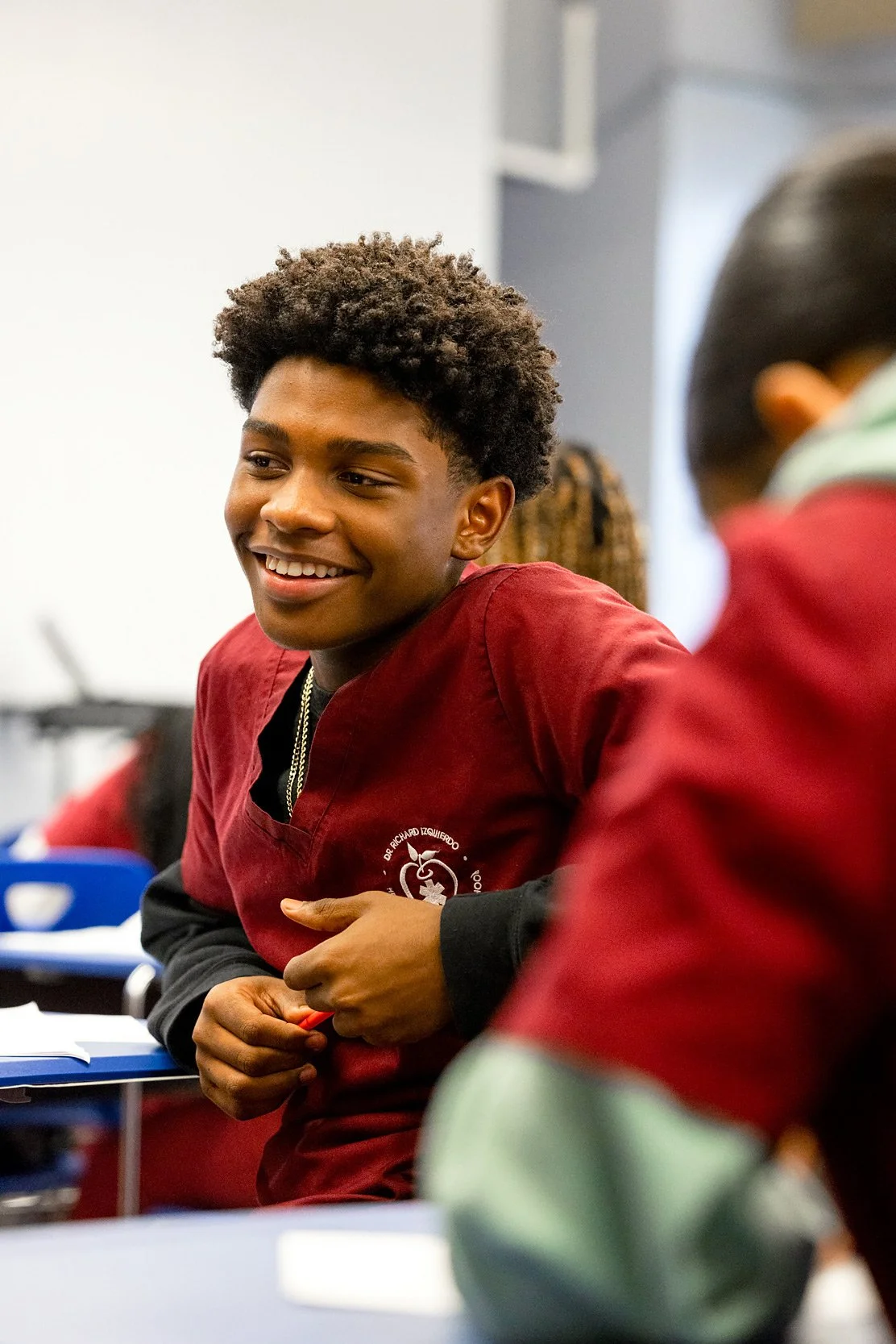 Student in red uniform smiling at his desk in classroom. 