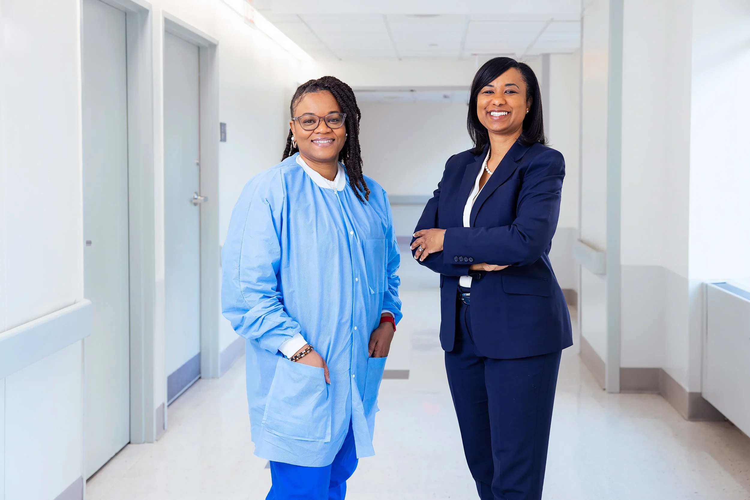 Female health professionals, one in hospital attire, one in a business suit posing in a bright hospital hallway.