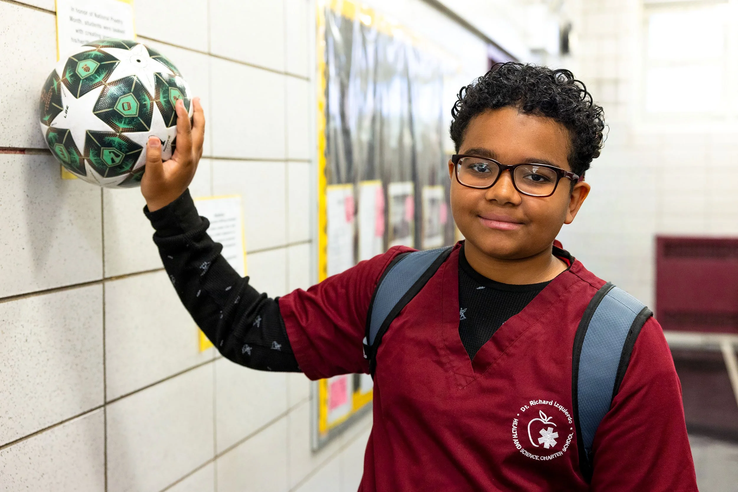 Student holding a soccer ball against a wall in a school hallway. 