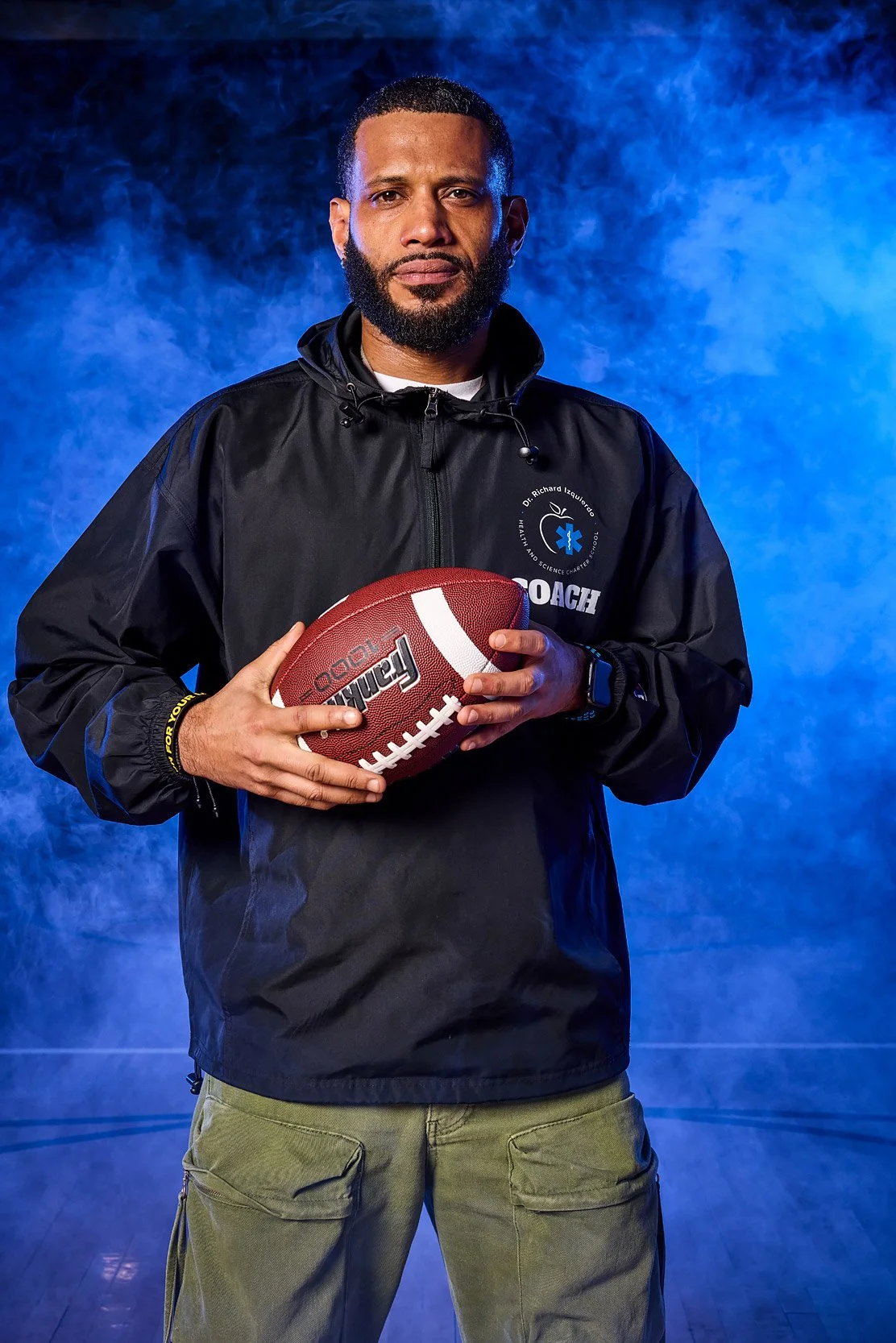Coach holding football, dramatic portrait against blue fog in a gym setting. 
