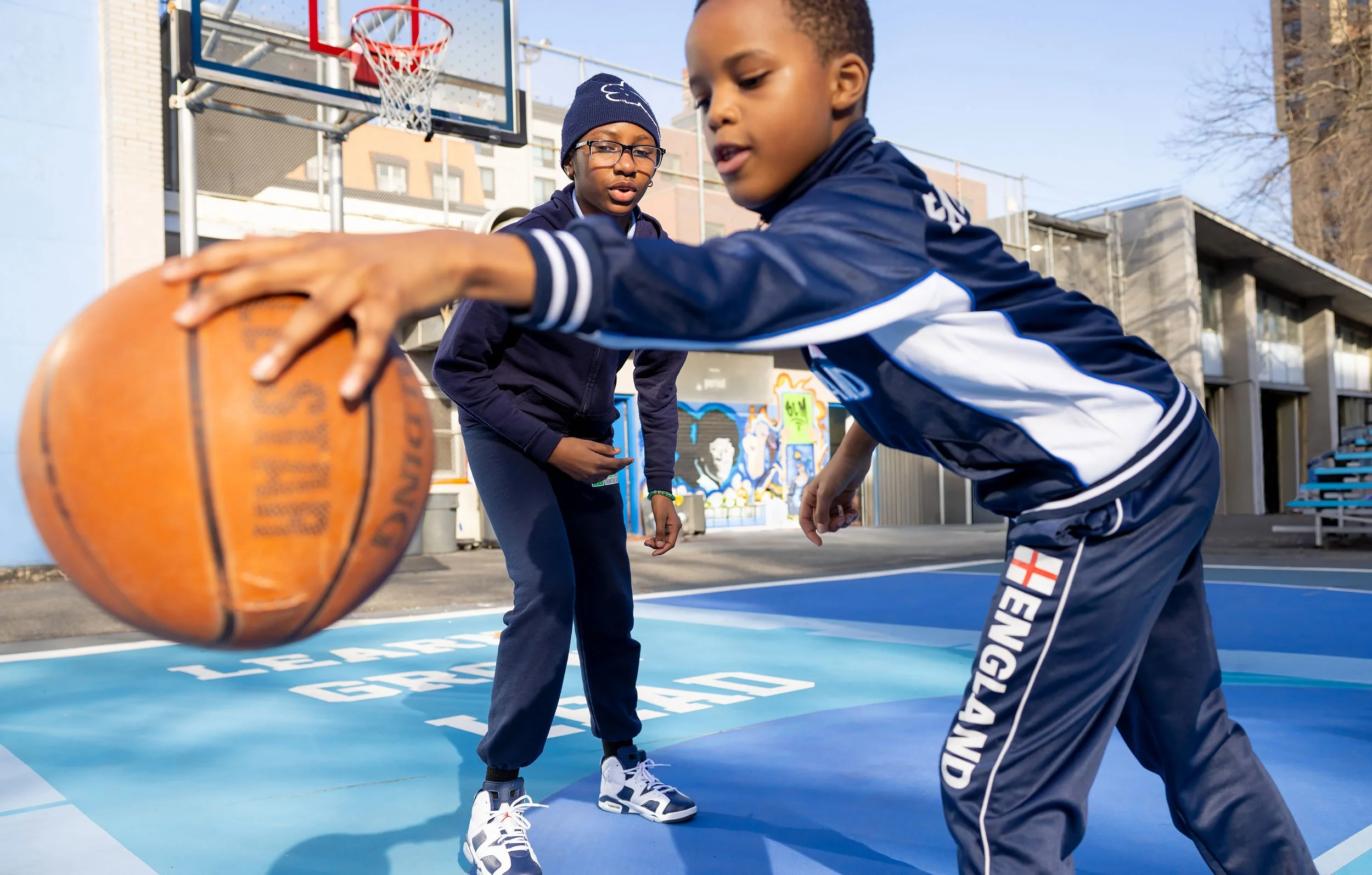 Two young students playing basketball outside at a court behind in their school yard.