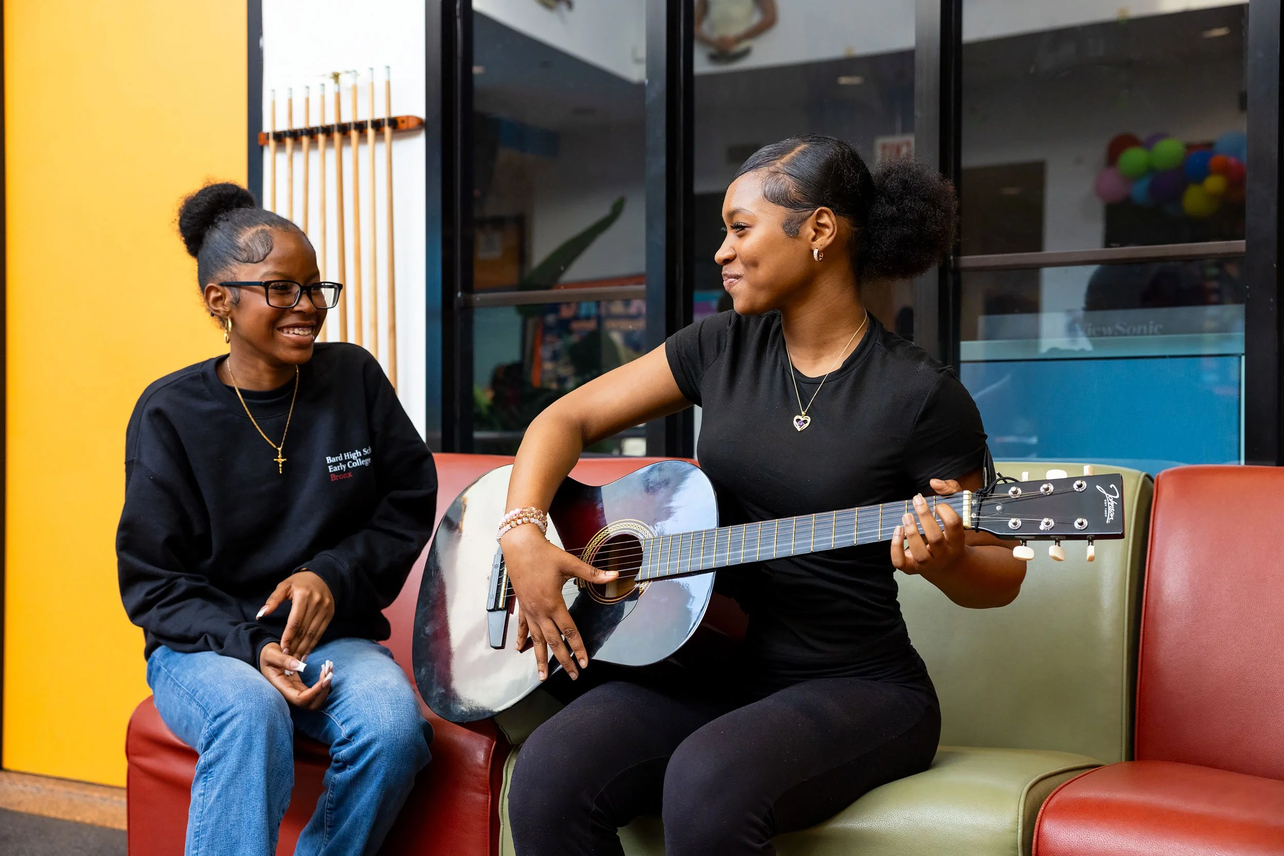 Student playing guitar with another student smiling and seated nearby in a school common area.
