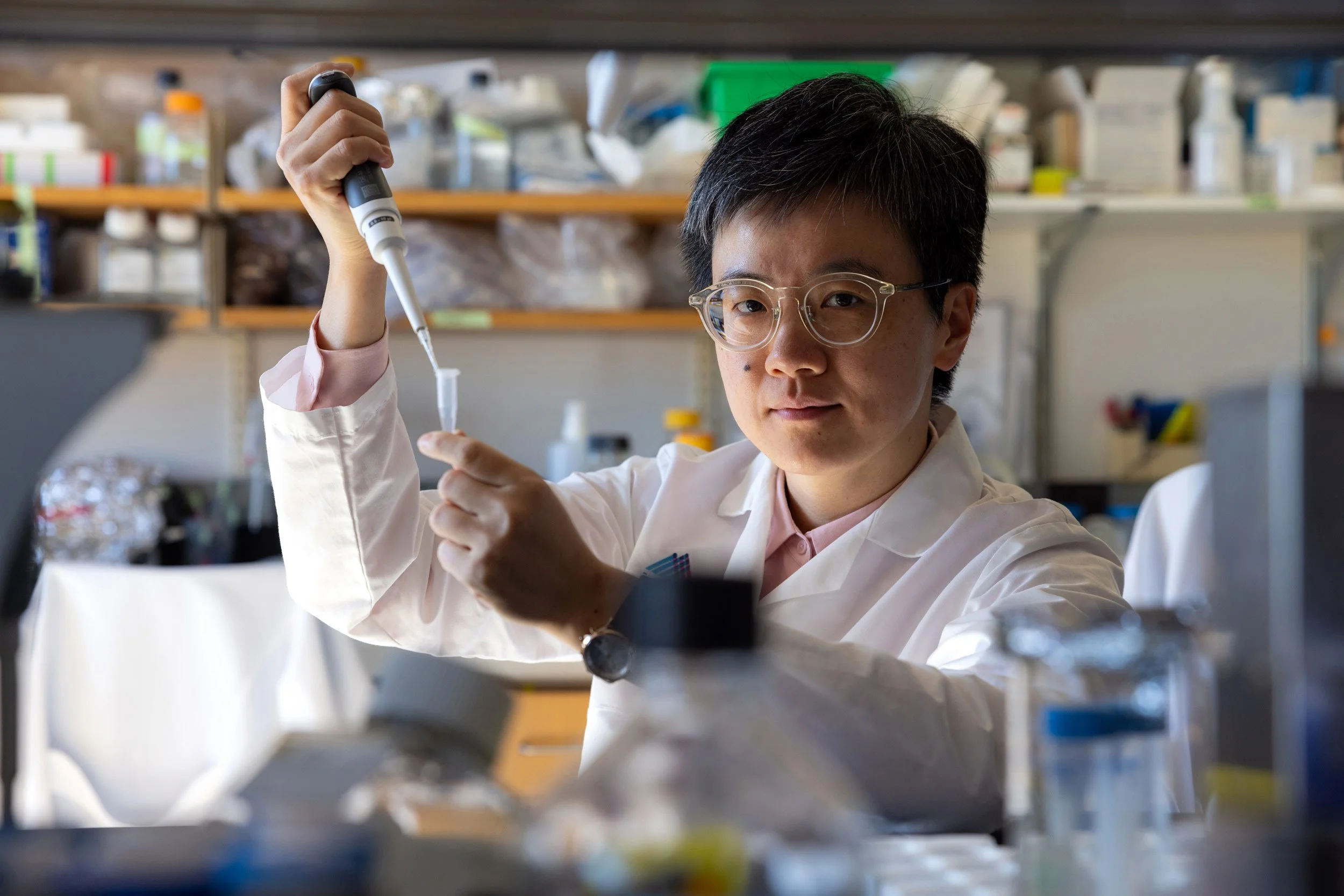 Female researcher with glasses and labcoat conducting a lab experiment with pipettes looking at the camera.