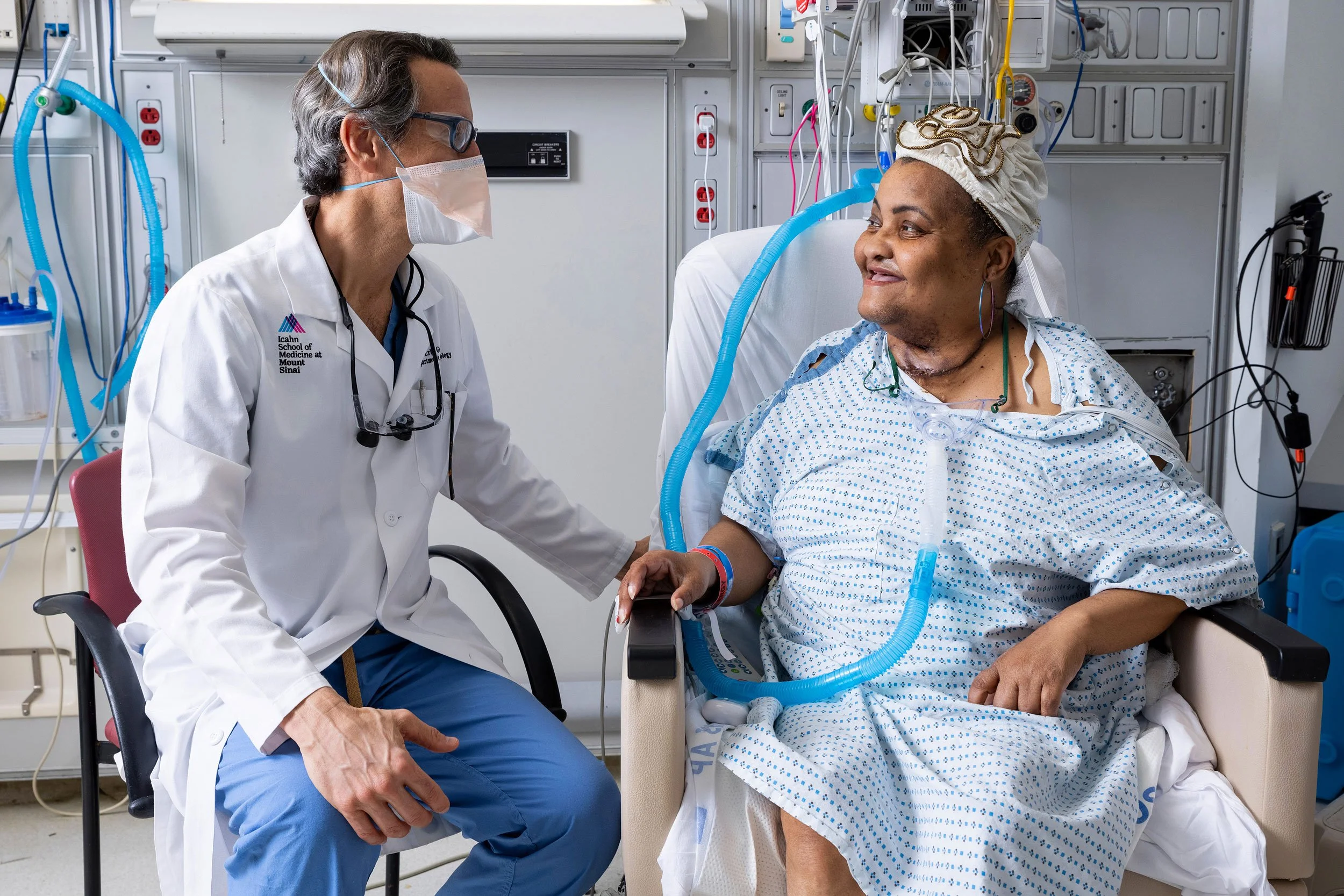 Doctor with surgical mask sitting beside a hospital bed talking with a smiling older patient.