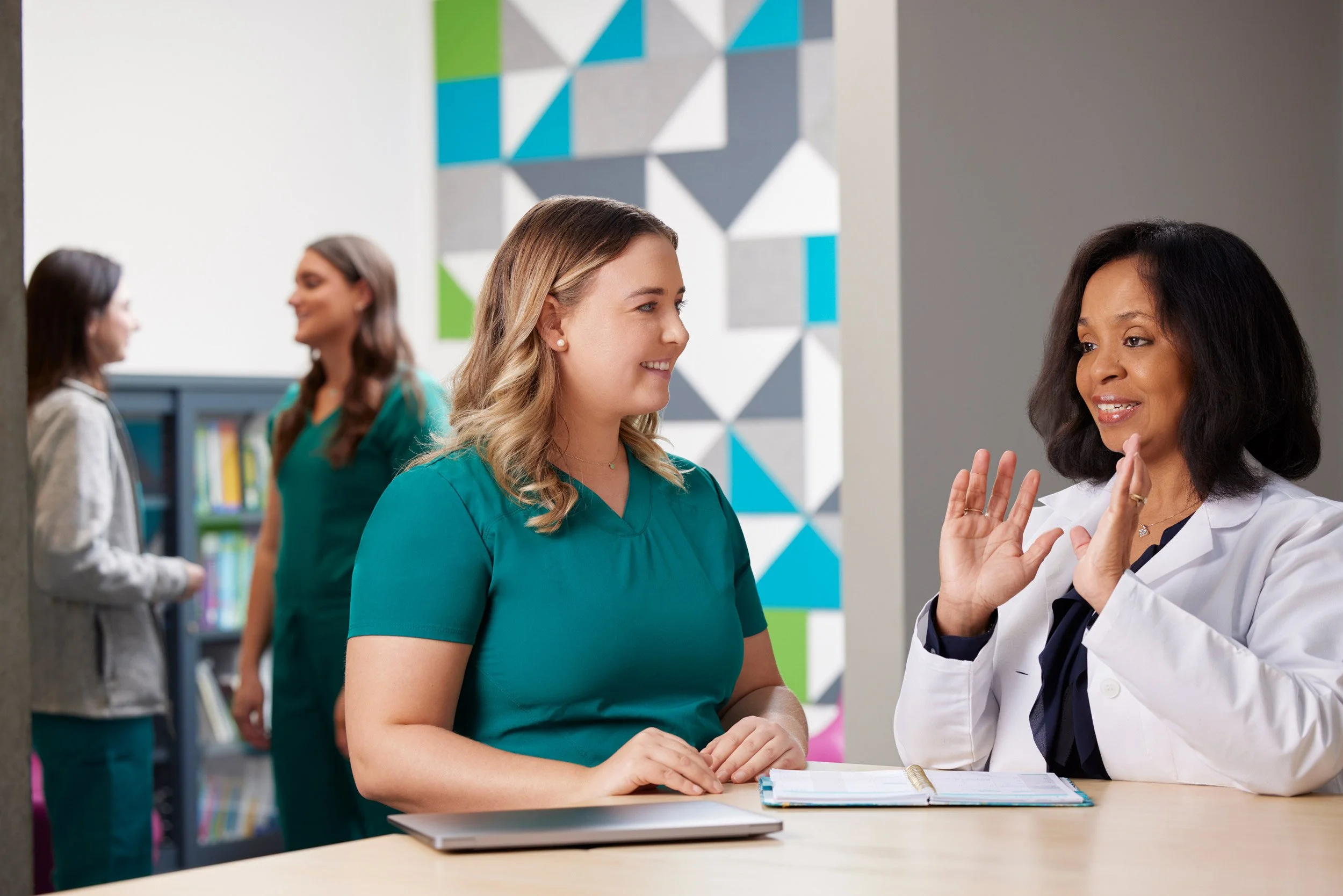 Two women in conversation at a clinic desk, one in green scrubs and one in a white lab coat.