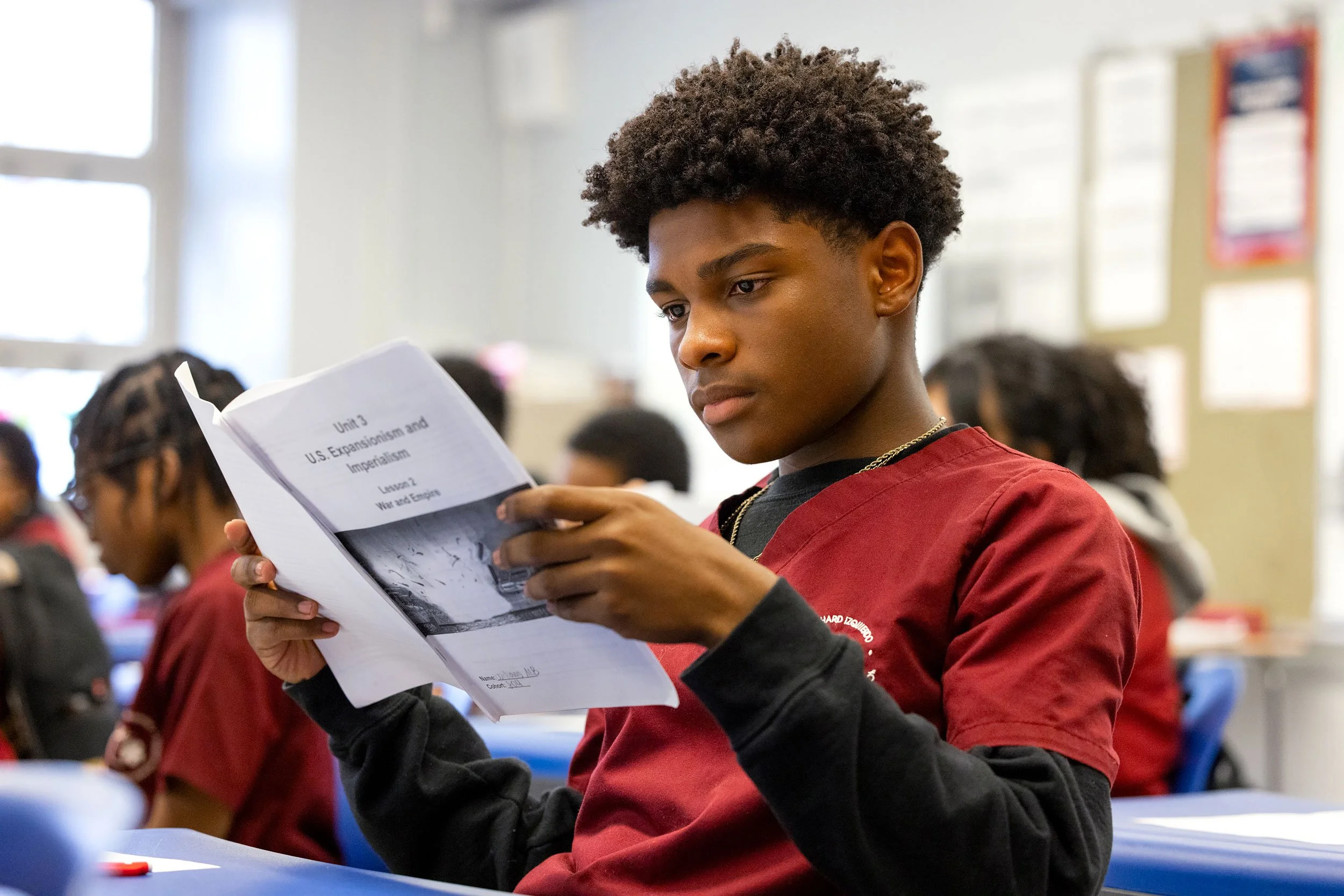Student reading a paper independently at desk in a quiet classroom moment.
