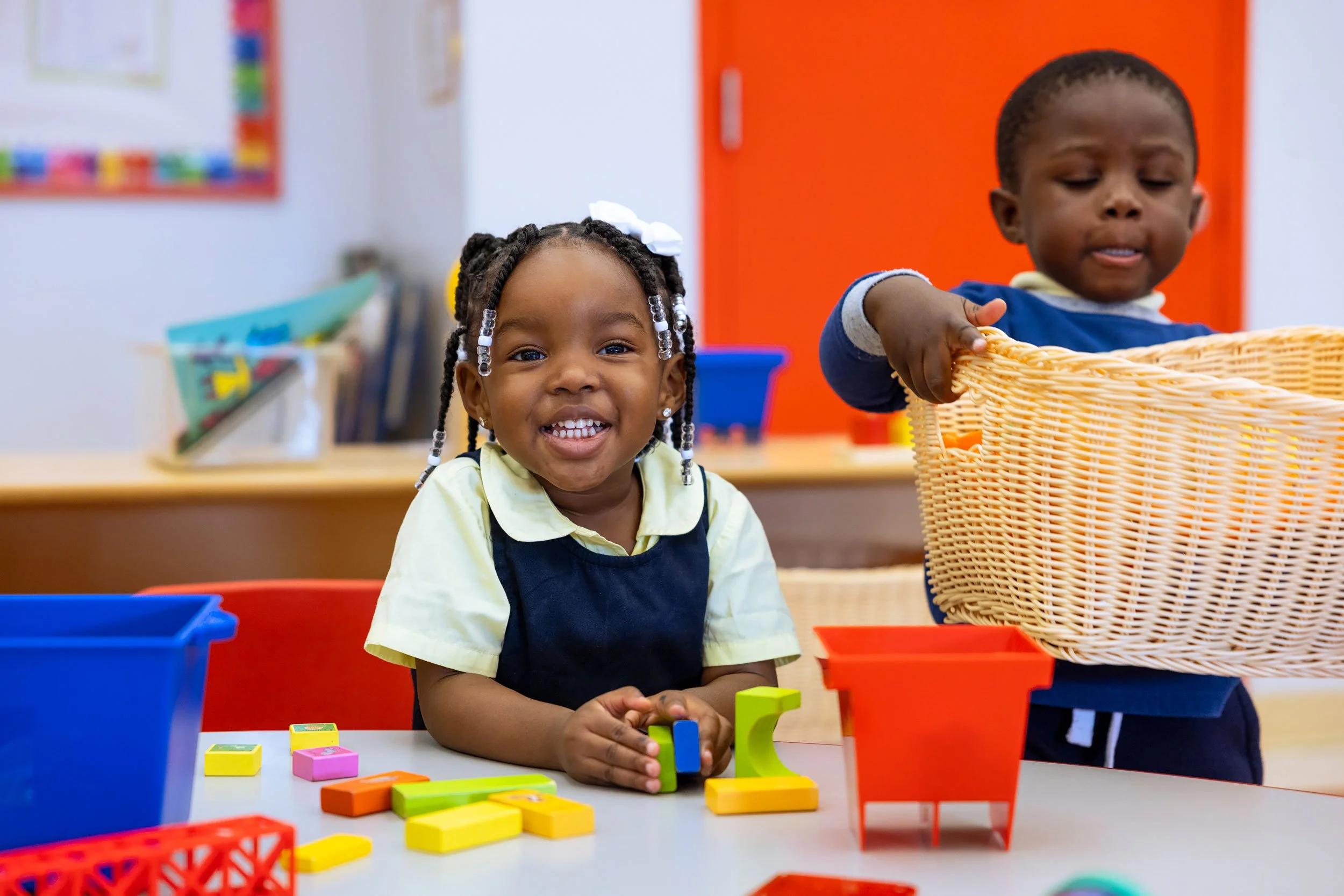 Young girl seated at table with colorful toys smiling while a young boy is carrying a woven basket.