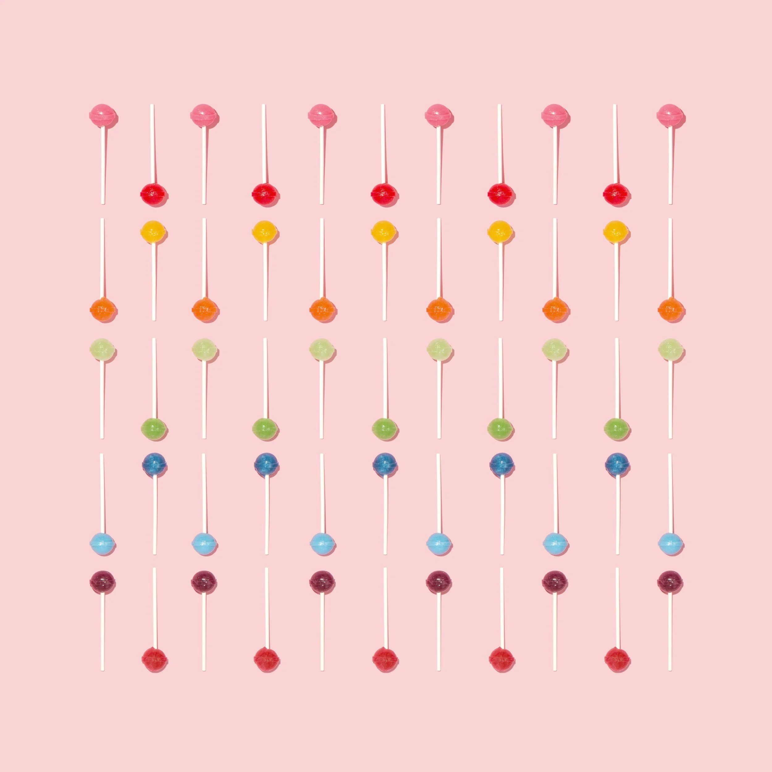 Mehrere bunte Lutscher mit weißen Stielen in Reihen auf rosa Hintergrund.