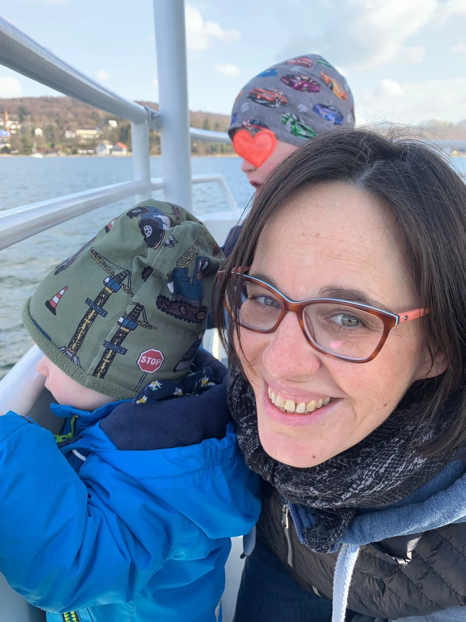 Frau mit Brille und Schal, lächelnd, begleitet zwei Kinder mit Hüten, auf einem Boot auf dem Wasser bei schönem Wetter.