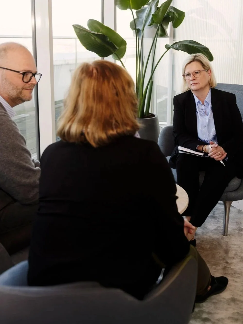 Tre personer sidder og taler i et moderne kontormiljø med store vinduer og en stort grøn plante.