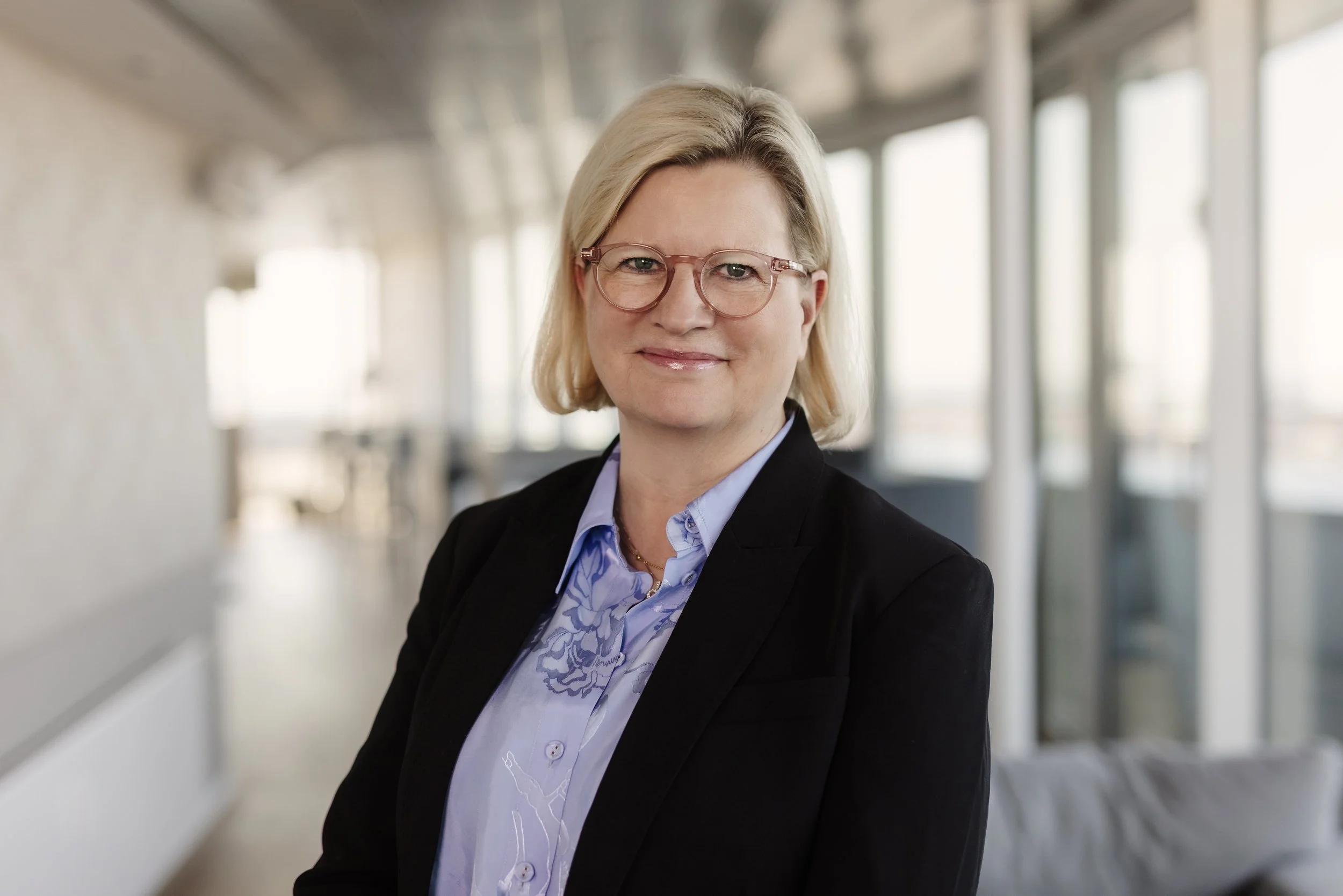 En kvinde med blondt hår, glasögon och dresseret tøj smiler i en moderne kontorbygning.