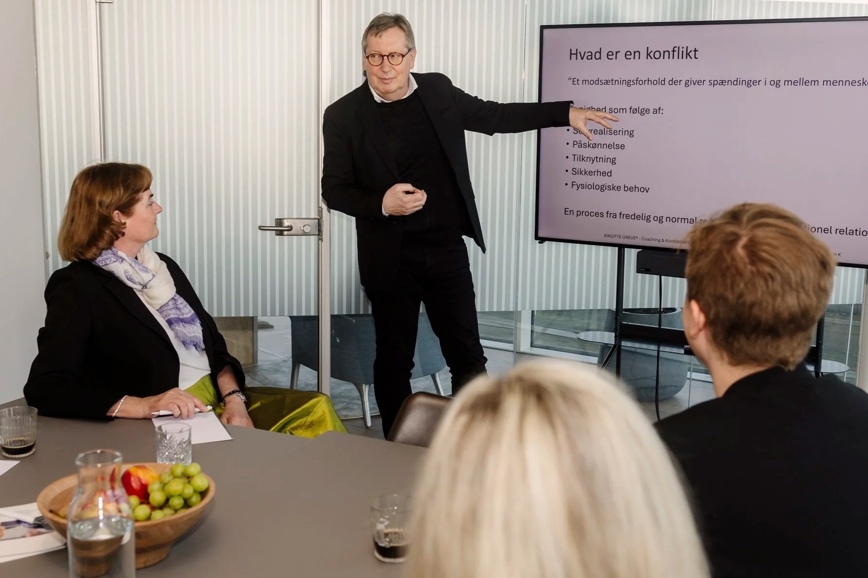 En mand holder en præsentation foran en skærm med teksten 'Hvad er en konflikt', mens en kvinde ser på ham. Der er flere personer, der lytter, og der er en skål med frugt på bordet.