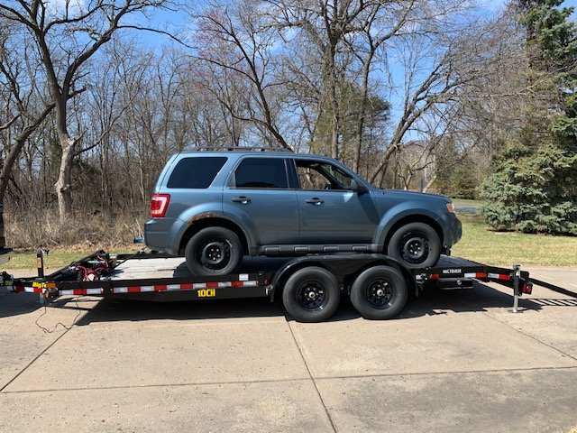 A gray SUV loaded on a flatbed trailer parked on a driveway, surrounded by trees and grass.