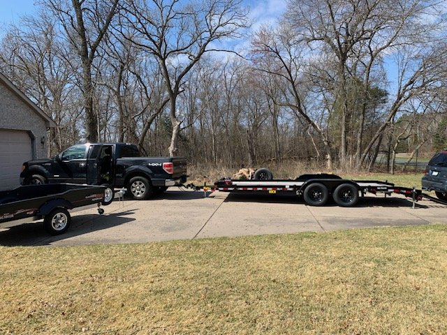 Pickup truck towing a flatbed trailer and a small camper parked on a driveway beside a garage, with trees and grass in the background.