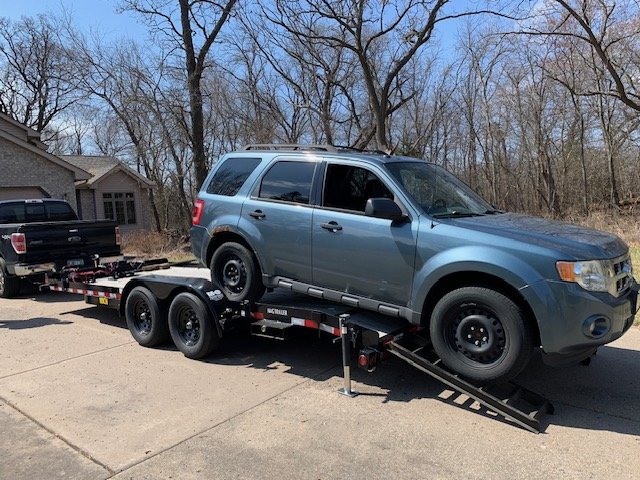 SUV being loaded onto a flatbed trailer attached to a pickup truck in a wooded area.