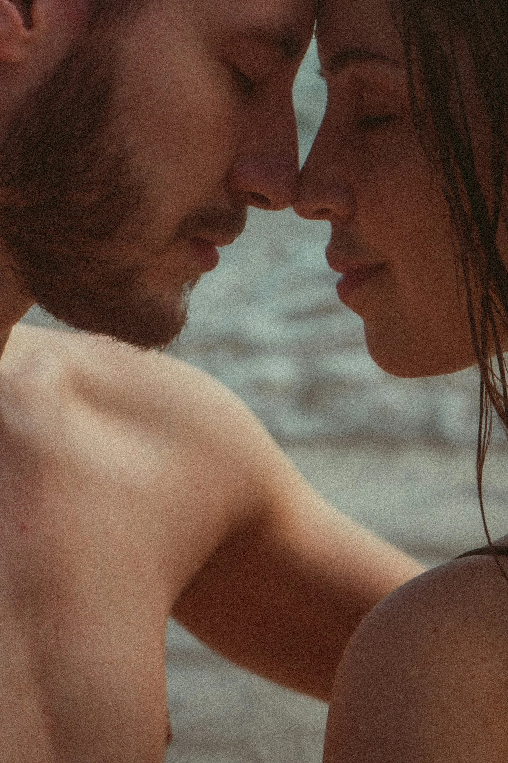 A close-up of a man and woman with their foreheads touching, eyes closed, by the water's edge.