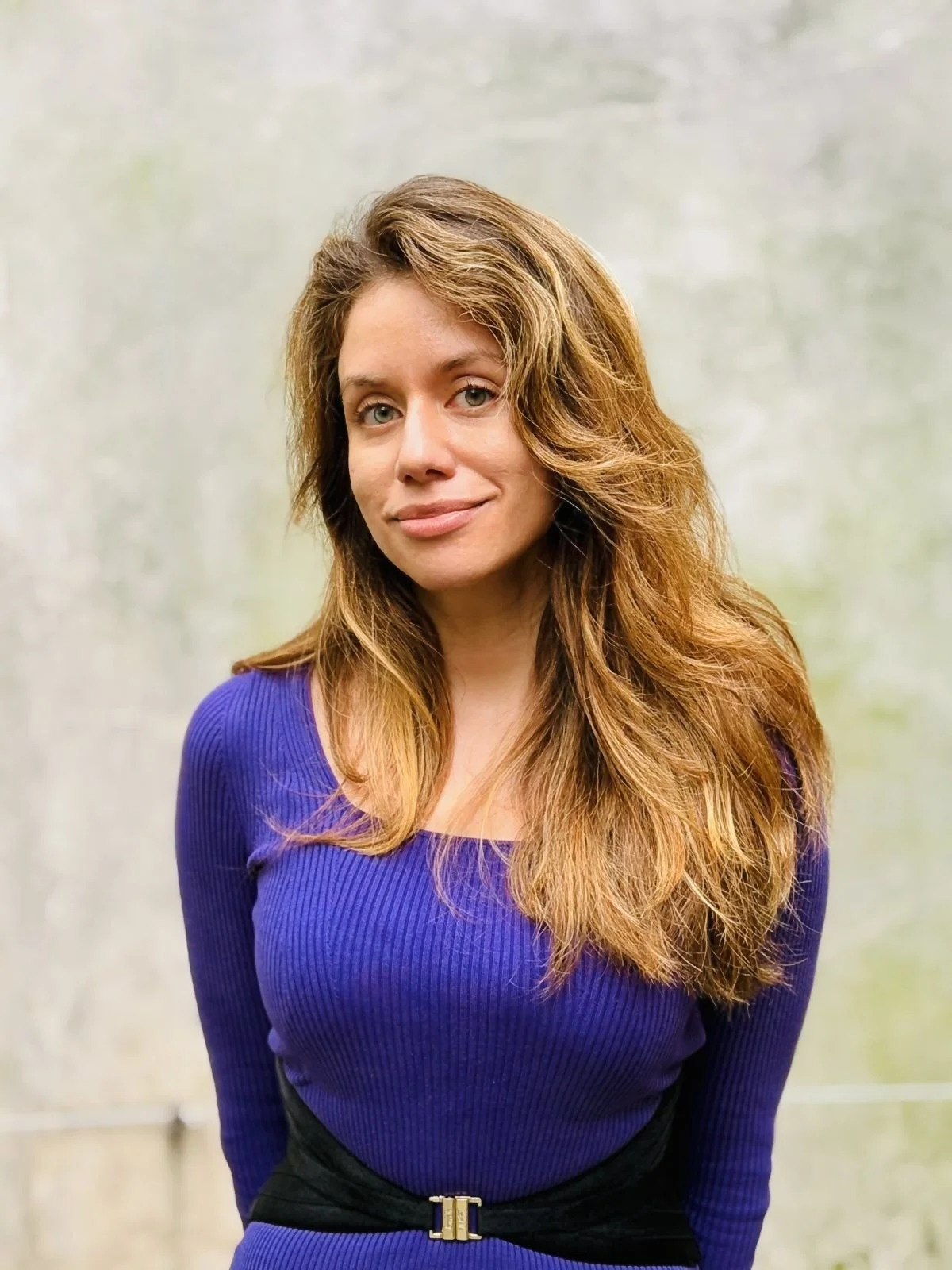 A woman with long, wavy, auburn hair wearing a purple, long-sleeved top with a black belt.