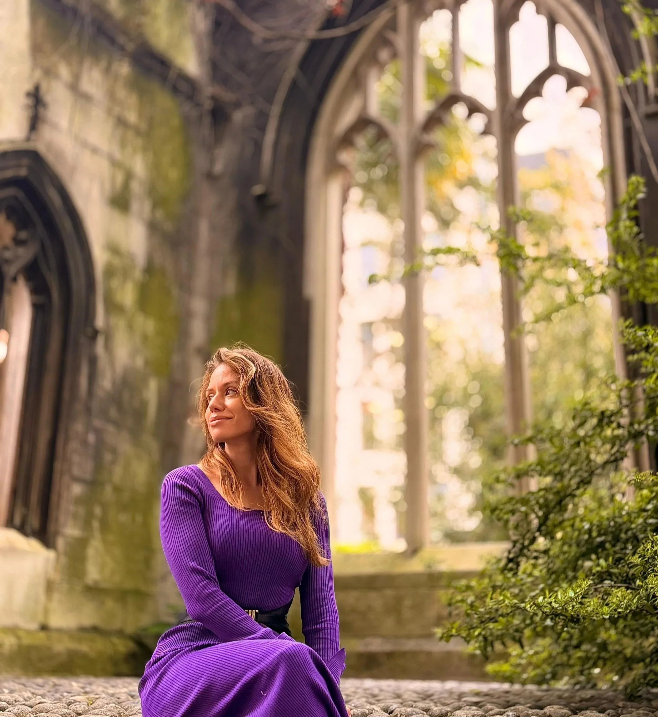 A woman with long, wavy hair wearing a purple long-sleeve top and a matching purple skirt sitting outdoors on a cobblestone ground near an old stone building with large, arched windows and greenery.