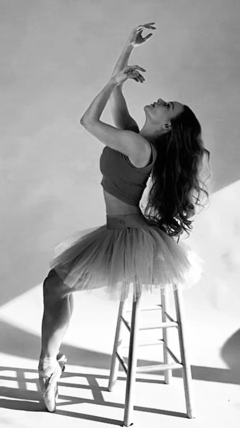 A woman dressed as a ballerina is sitting on a wooden stool, wearing a tulle tutu, pointe shoes, and a fitted top, with her arms raised artistically and head tilted back, creating a graceful pose.