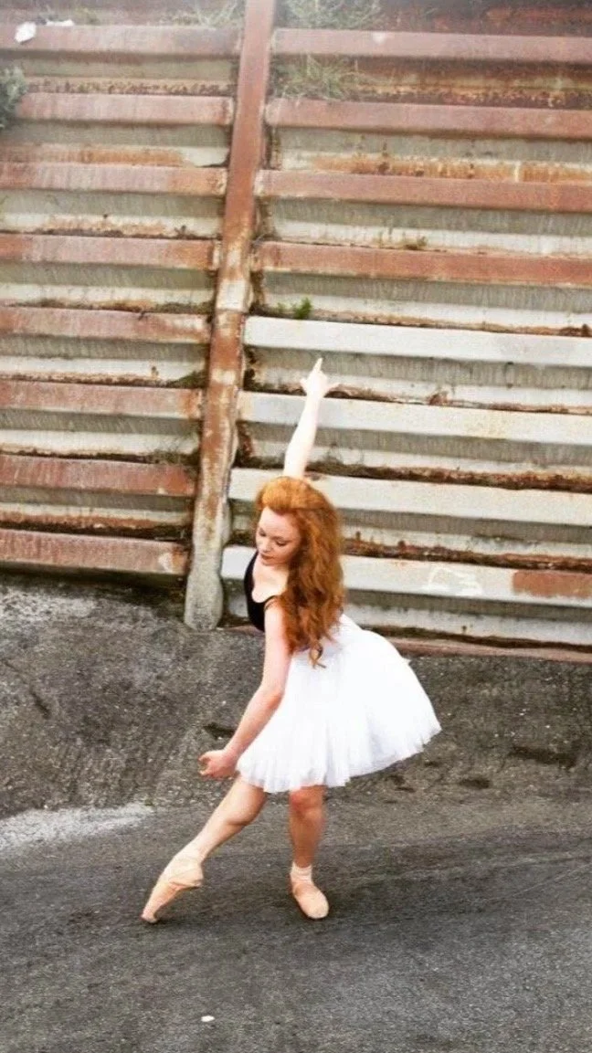 A ballerina with red hair in a tutu and pointe shoes posing on an outdoor street, with a weathered wooden fence in the background.