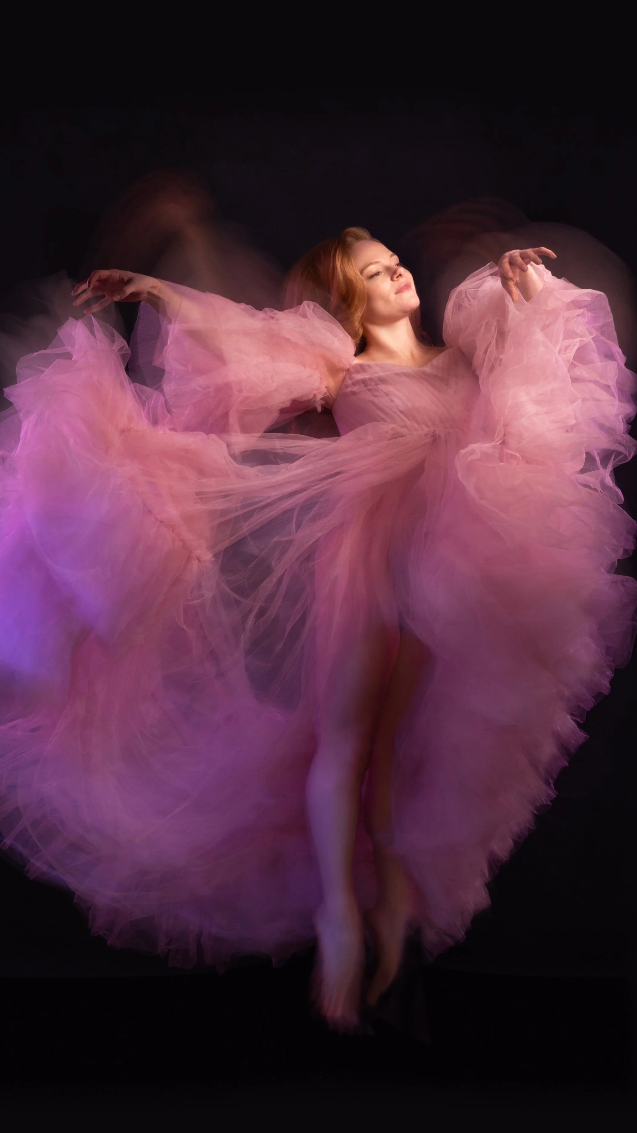 A woman in a flowing pink tulle dress is lying on a black background with her arms raised and her eyes closed.