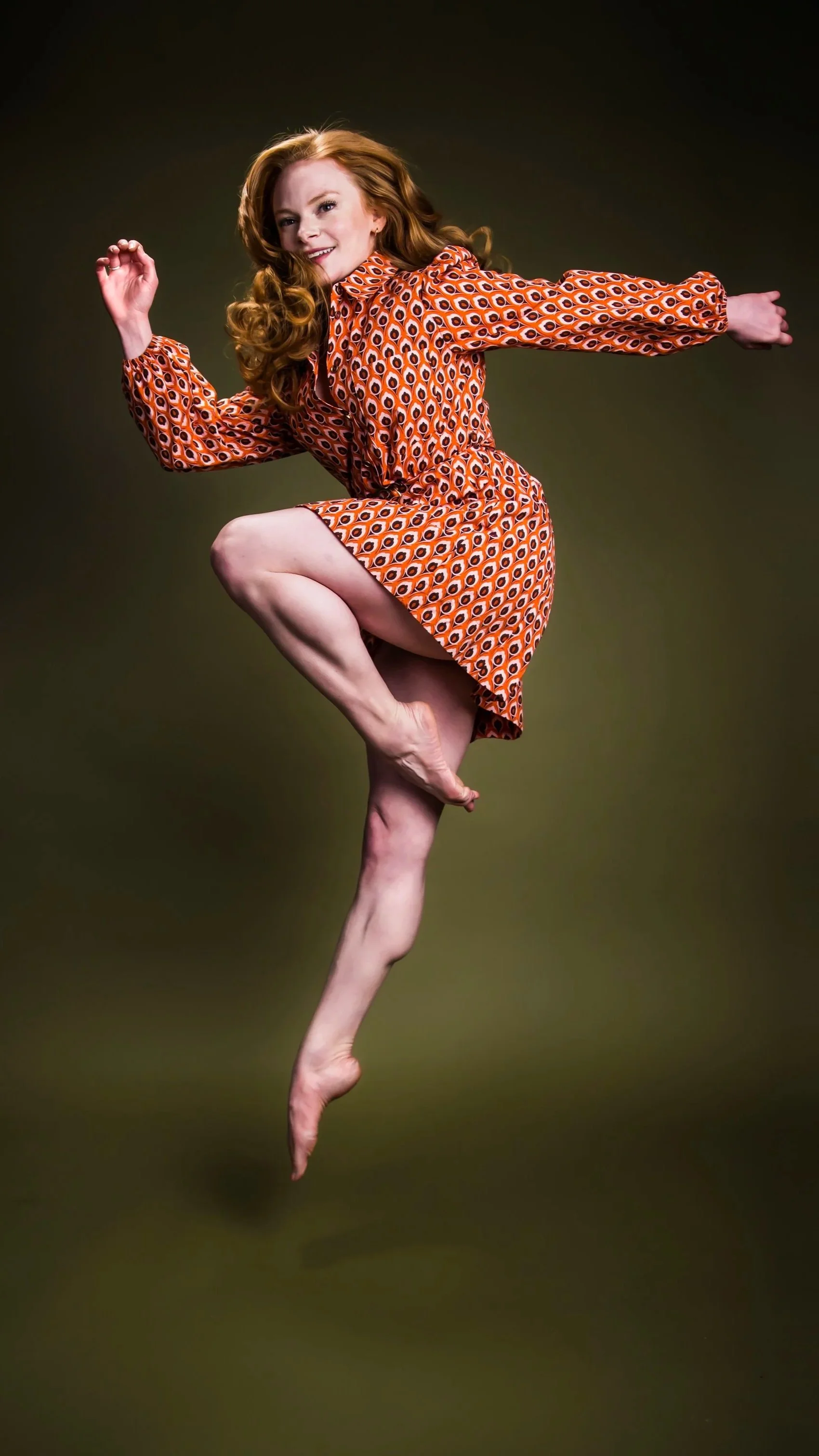 A woman with long red hair in a patterned orange dress is balancing on one leg with her other leg bent and raised, and her arm extended forward in a dynamic pose.