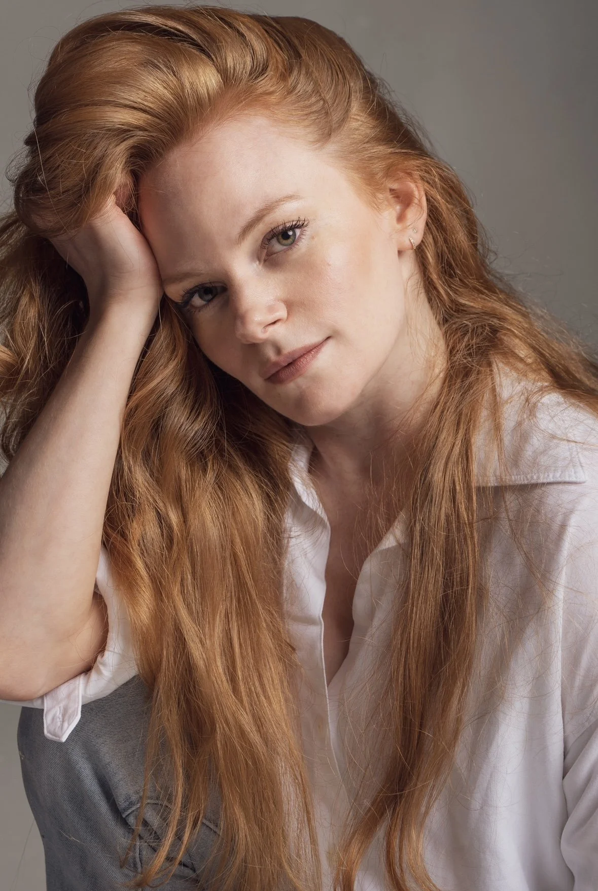 A woman with long, wavy red hair and blue eyes, wearing a white shirt, resting her head on her hand, looking at the camera with a slight smile.