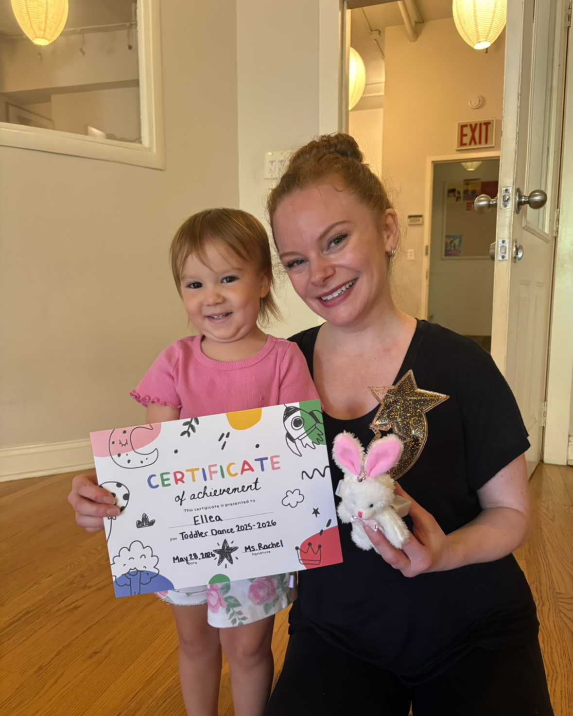 A smiling young woman and a little girl standing together indoors, holding a certificate of achievement. The woman has red hair styled in a bun and wears a black shirt decorated with a star and bunny plush; the girl wears a pink shirt and floral shorts. There are paper lanterns hanging from the ceiling in the background.