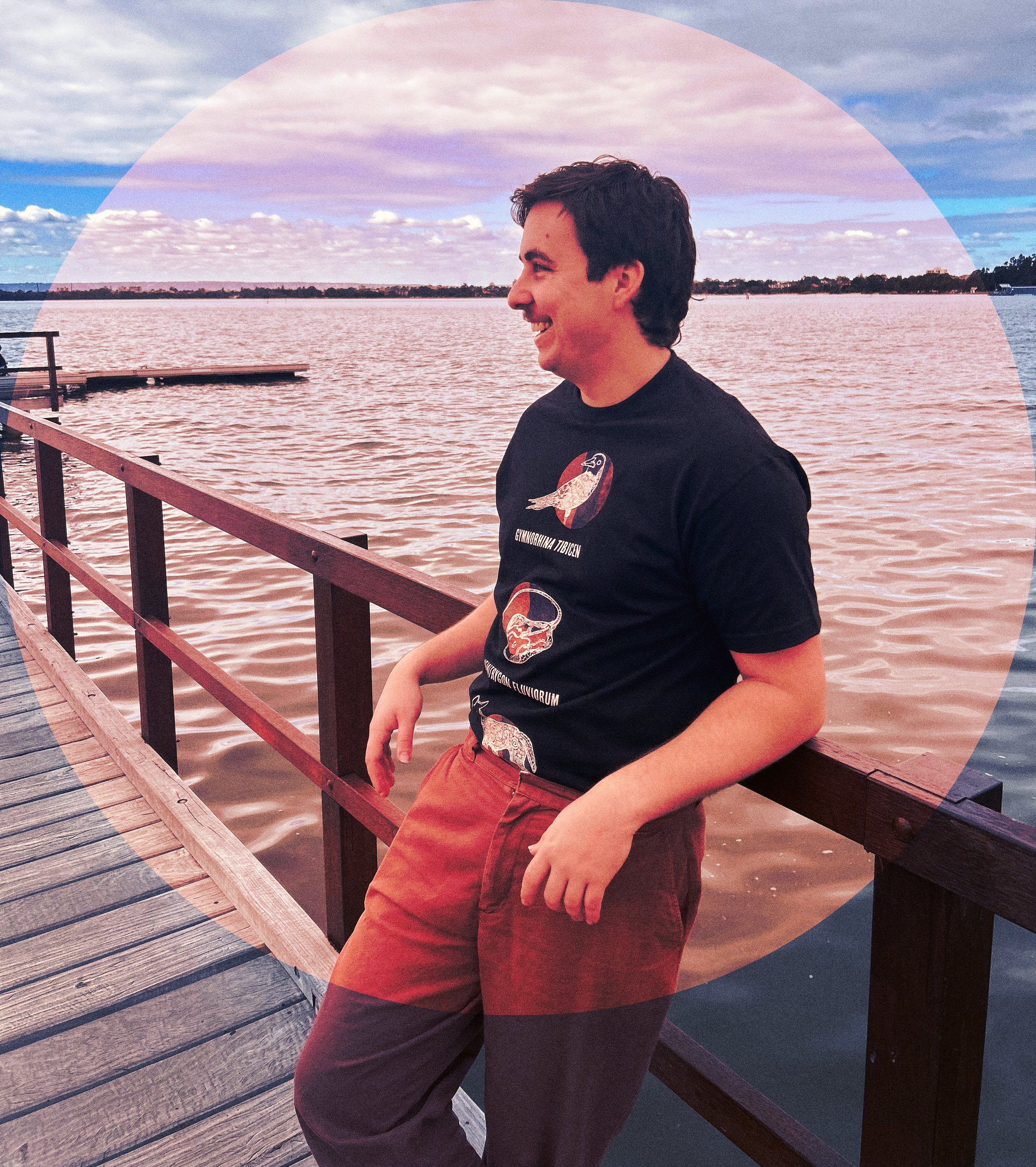 A young Indigenous man leaning on a pier railing by a lake, smiling and looking to the side, with an Indigenous shirt