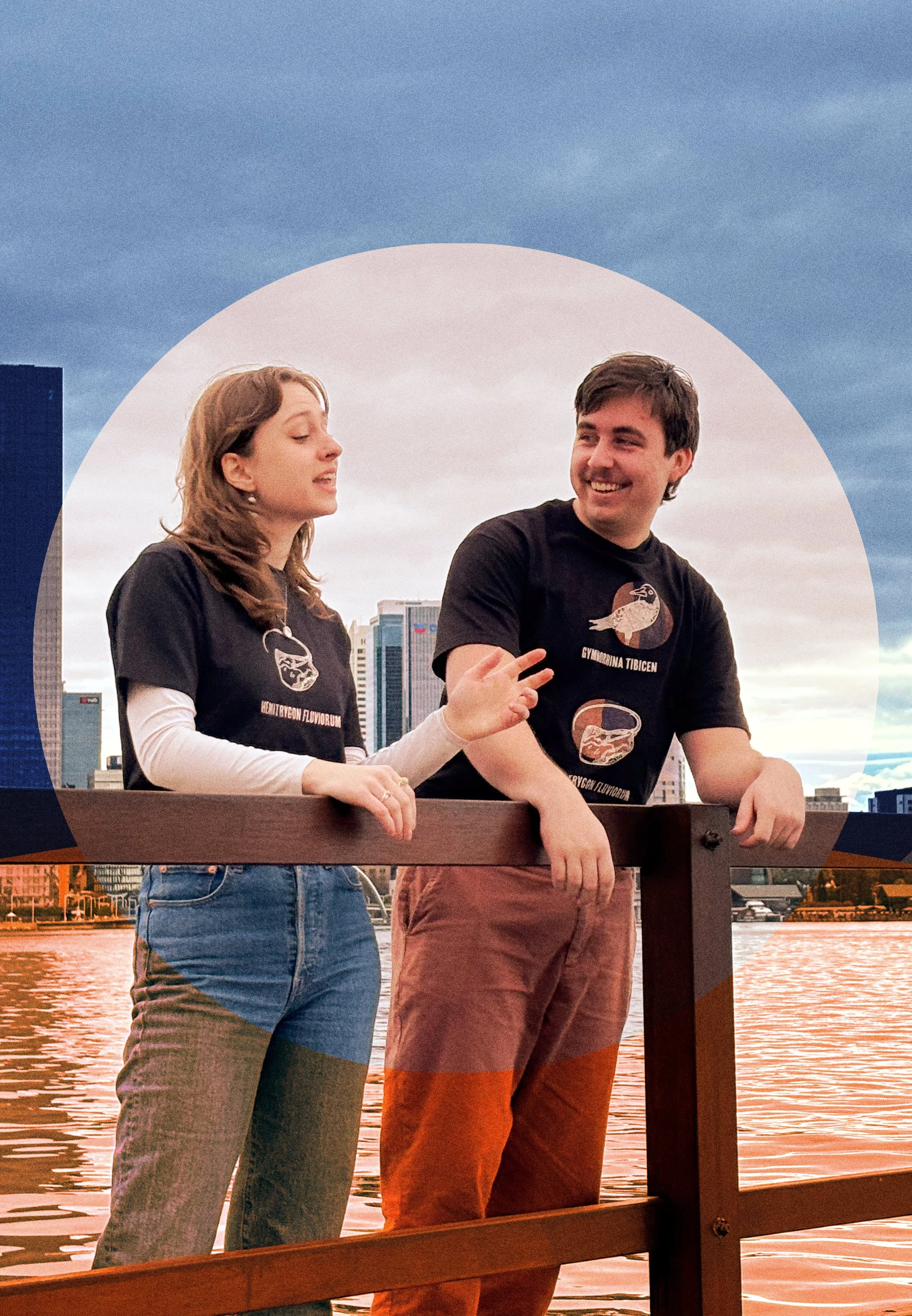 A woman and a man are standing on a dock by the water, engaged in conversation, with Indigenous designed shirts, with a city skyline and cloudy sky in the background.
