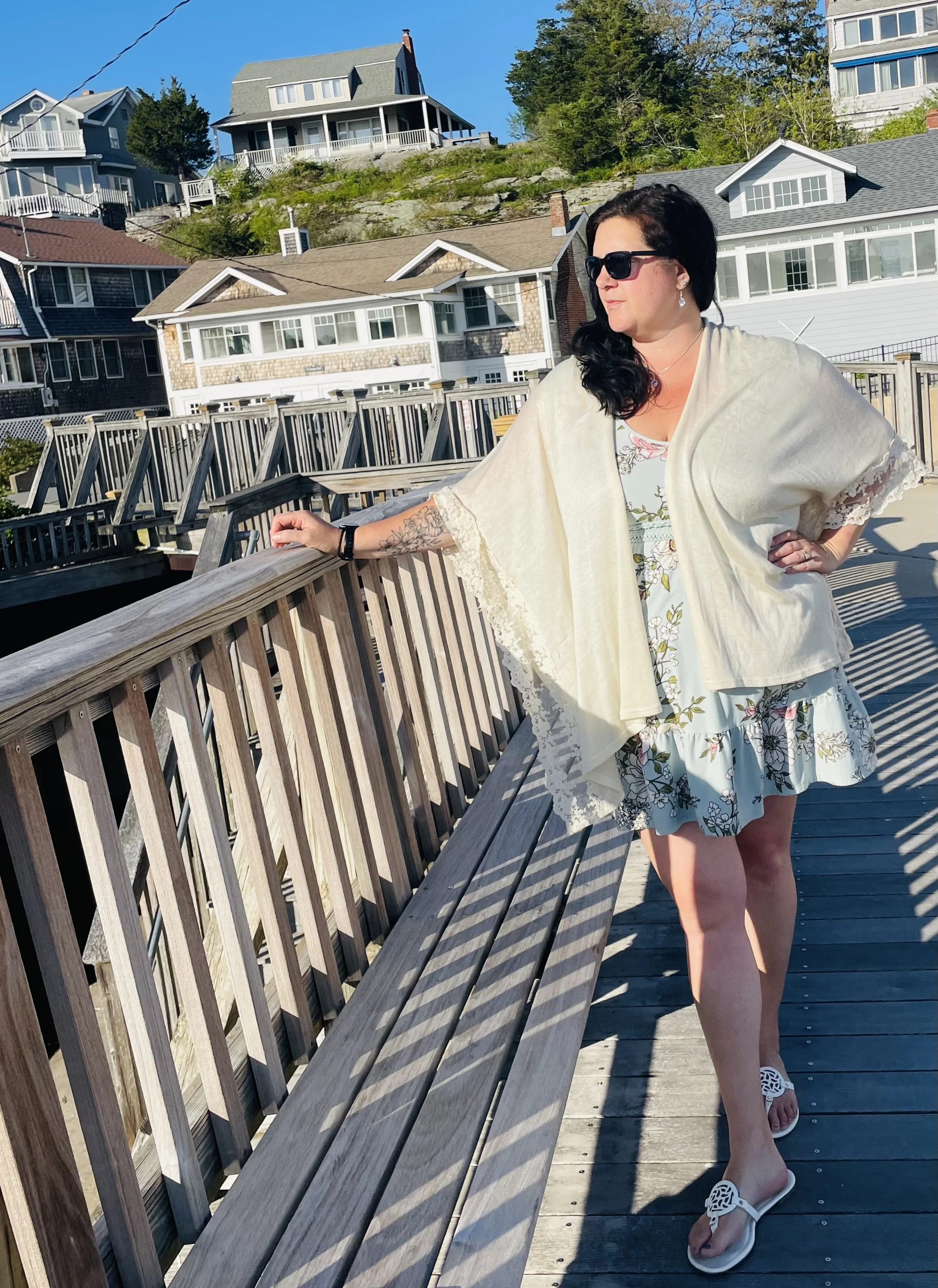 Woman standing on a wooden boardwalk near the water, wearing sunglasses, a floral dress, a light shawl, and white sandals, with houses on a hill in the background under a clear blue sky.