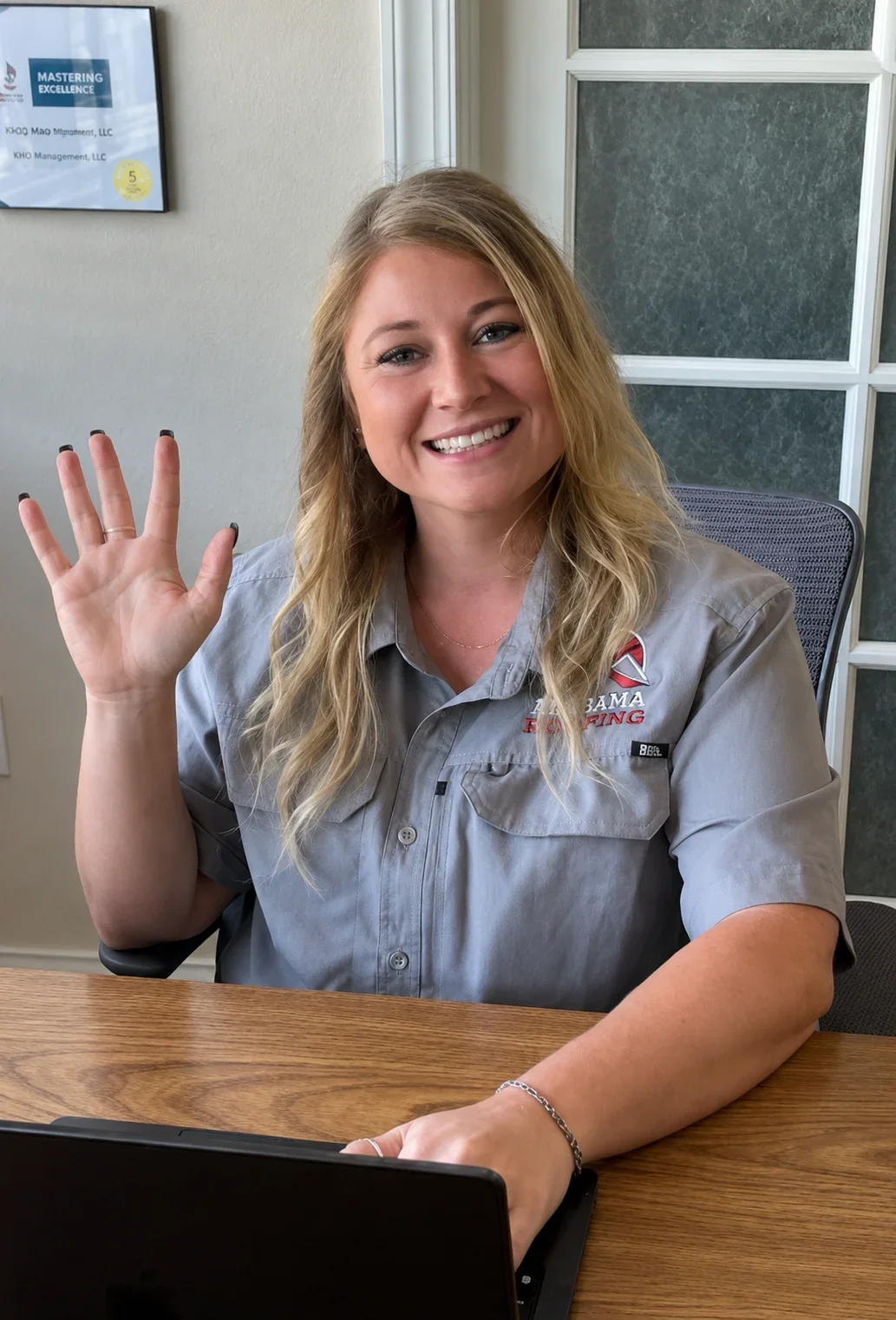 Lucy, Office Assistant for Craig Fass Roofing, smiling and waving at her desk in the Homosassa office.