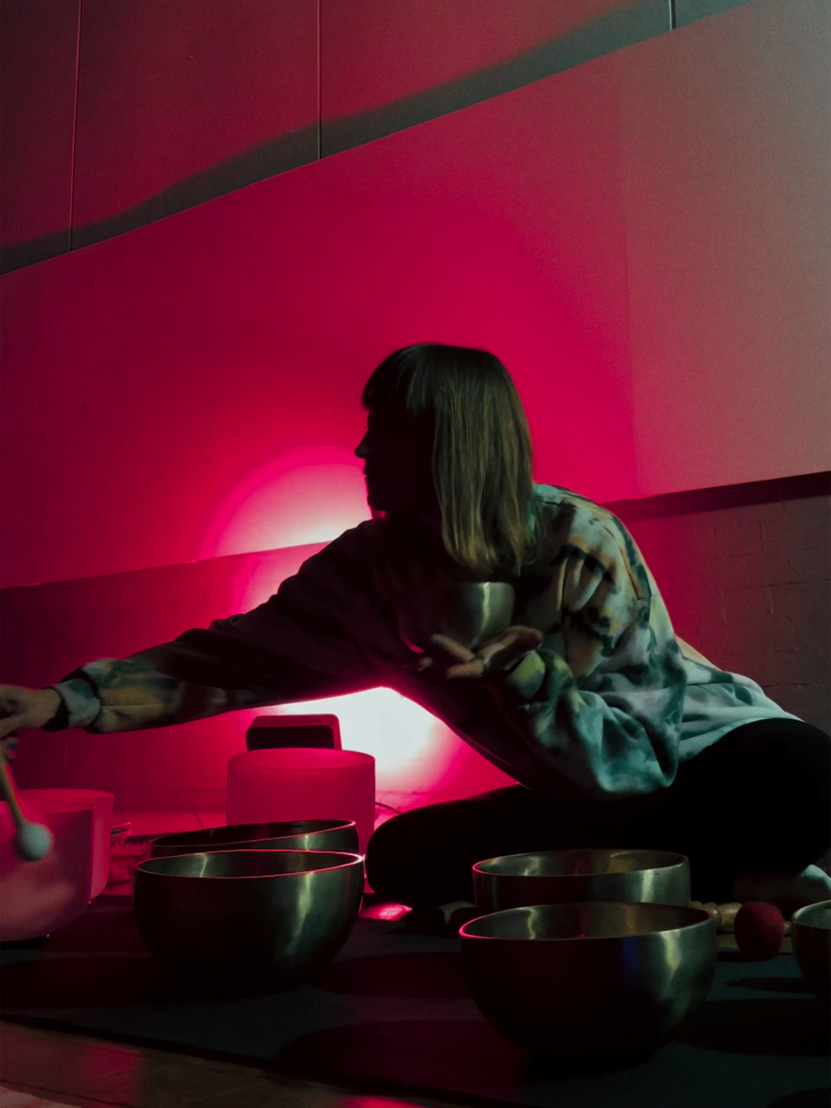 A person with shoulder-length hair is sitting cross-legged on the floor, reaching for items in front of them, with a pinkish-red light illuminating the background and casting a glow on their face and body. Several bowls and objects are placed on the floor around them.