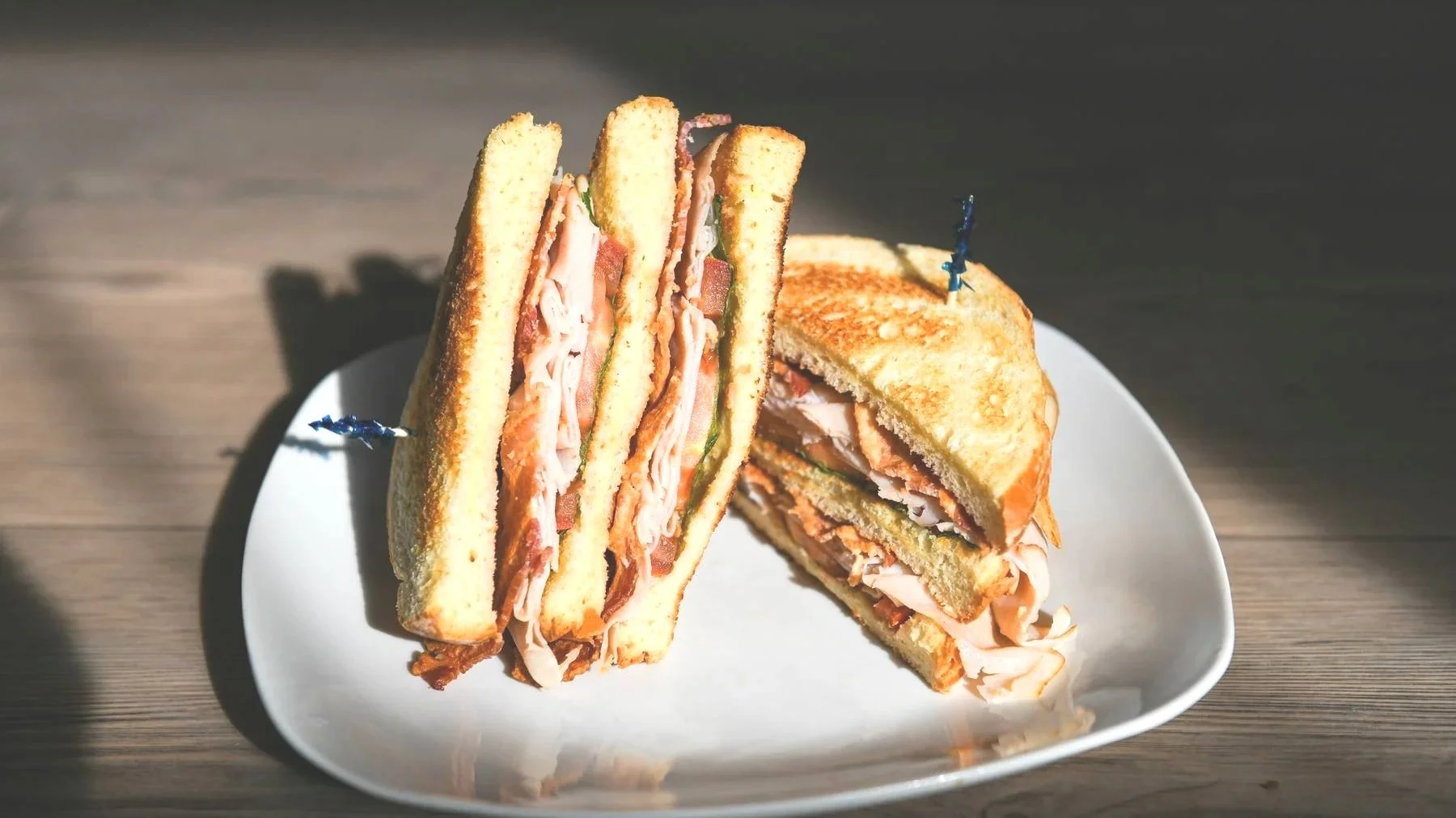 A plate of three toasted sandwiches with turkey and bacon, cut into halves, arranged on a white plate on a wooden surface with sunlight.