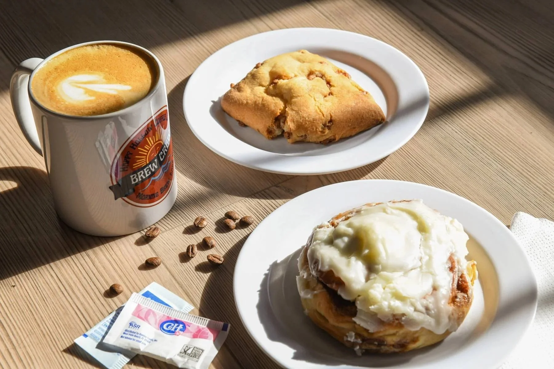 A breakfast meal with a cup of coffee, a cinnamon roll with icing, and a cinnamon scone on plates, with coffee beans and sugar packets on a wooden table.
