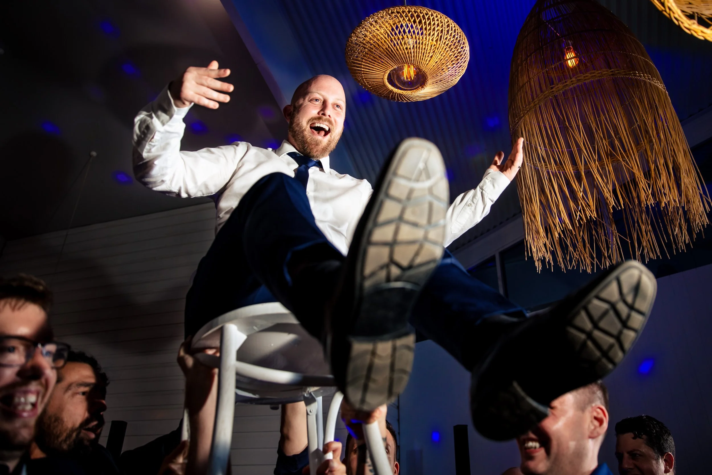 A groom being lifted in a chair above a crowd, smiling and waving. Taken at Ancora, by Hunter Valley & Port Stephens wedding photographers, Lynn & Justin.