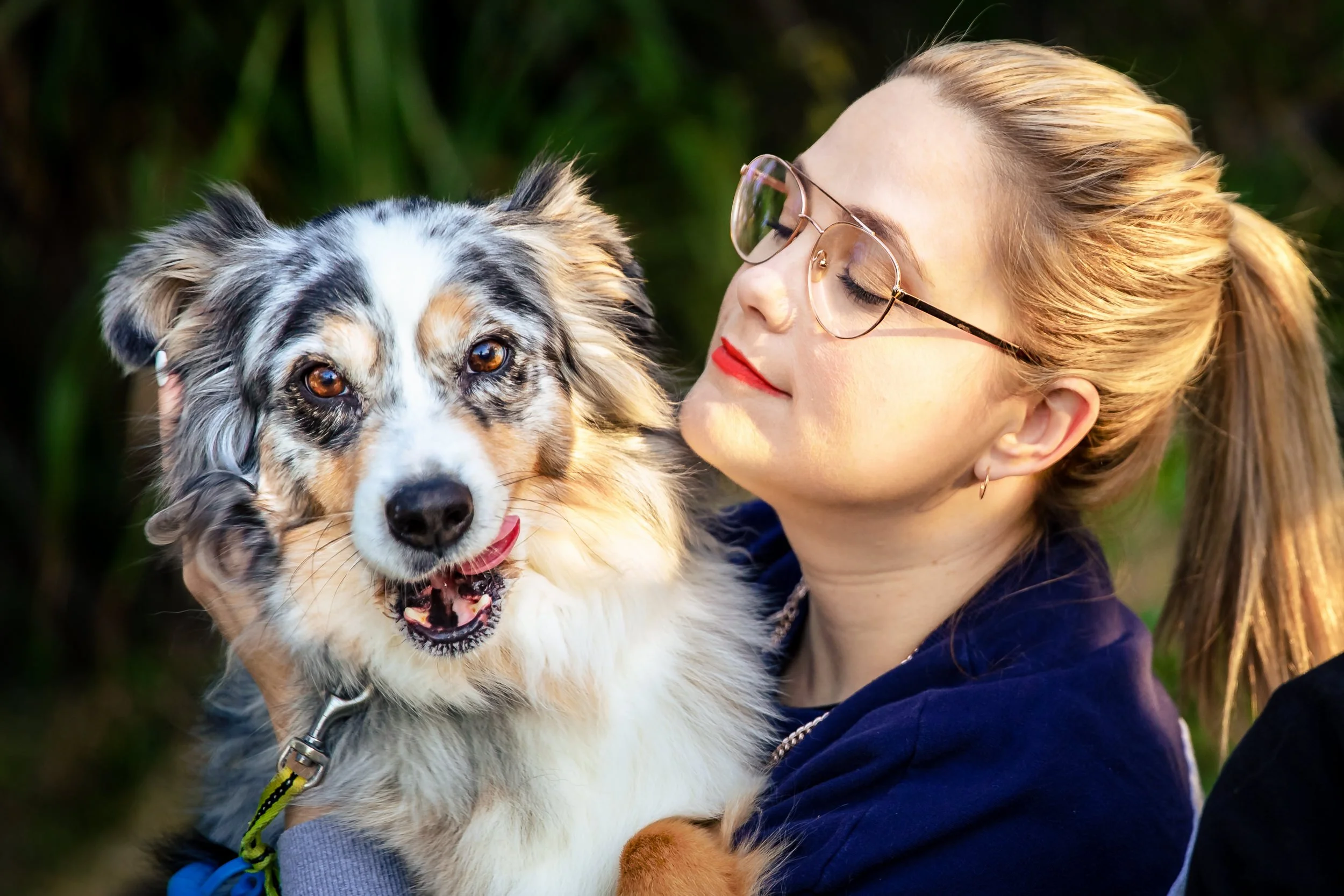 A woman with blonde hair in a ponytail, wearing glasses and red lipstick, holding a tricolor Australian Shepherd dog with blue eyes, outdoors with green foliage in the background in Singleton, NSW.