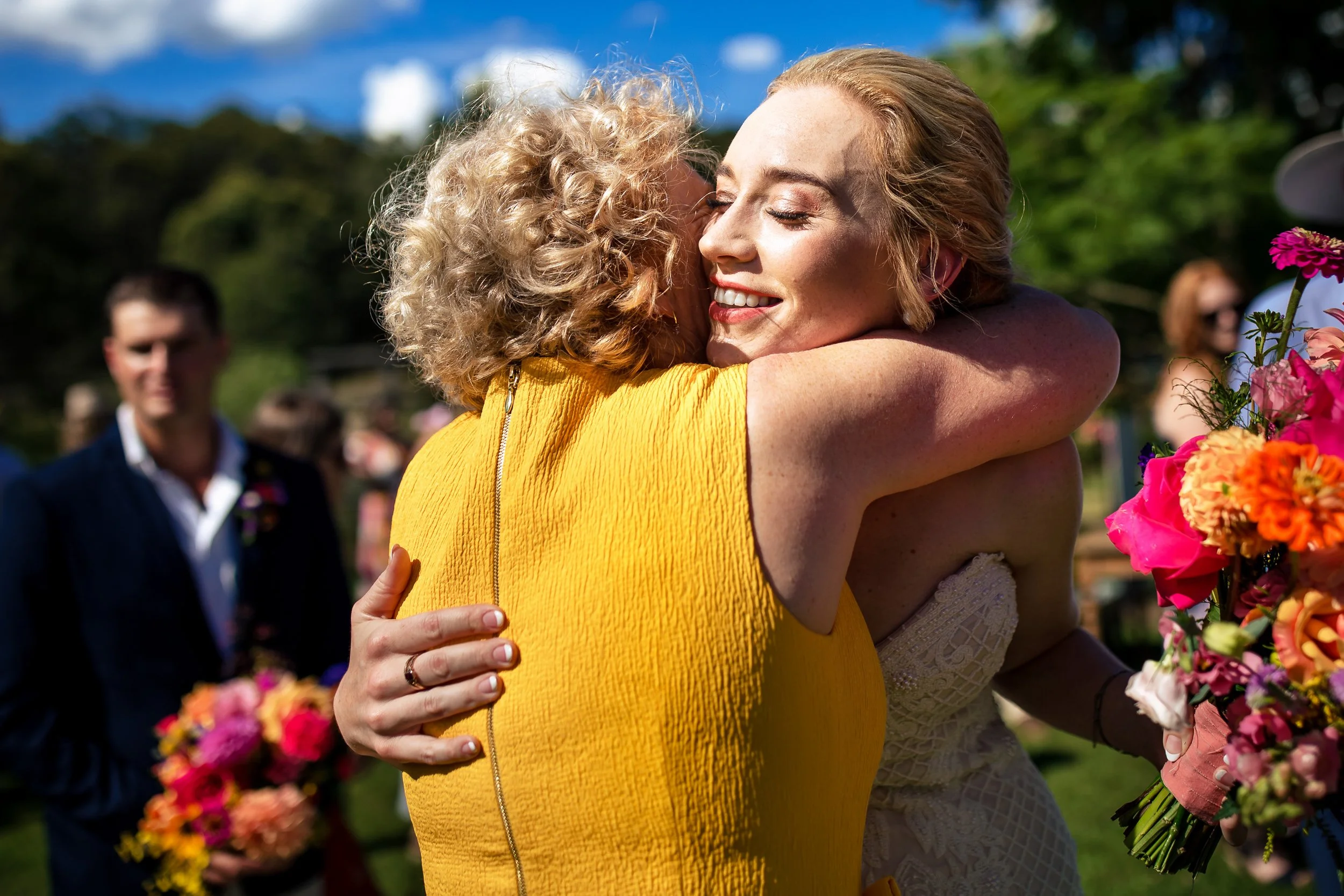 A bride hugging her Mum after a wedding ceremony in Singleton, NSW, with vibrant flowers in the foreground and guests in the background.