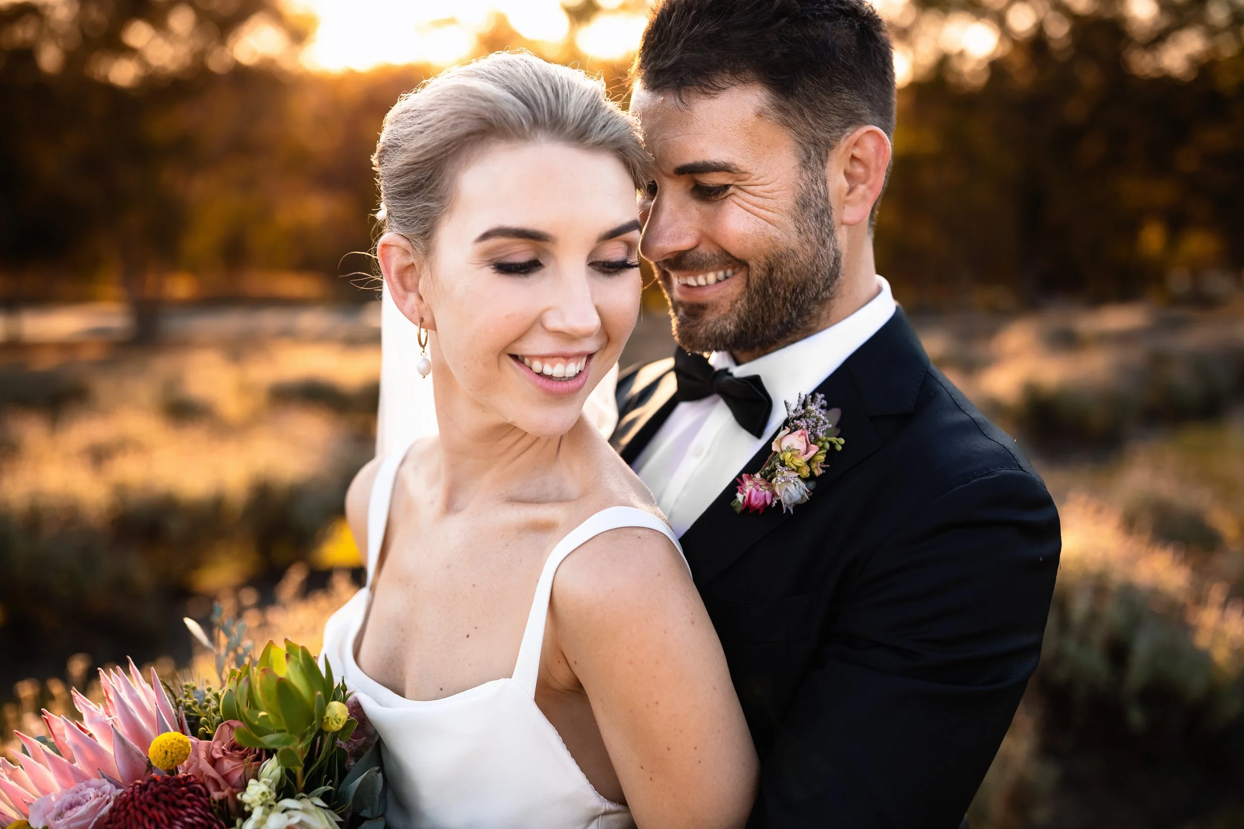 A happy couple on their wedding day, embracing outdoors during sunset. The bride holds a bouquet of pink and green flowers, and both are dressed in wedding attire. Captured at by Hunter Valley wedding photographers, Lynn & Justin.