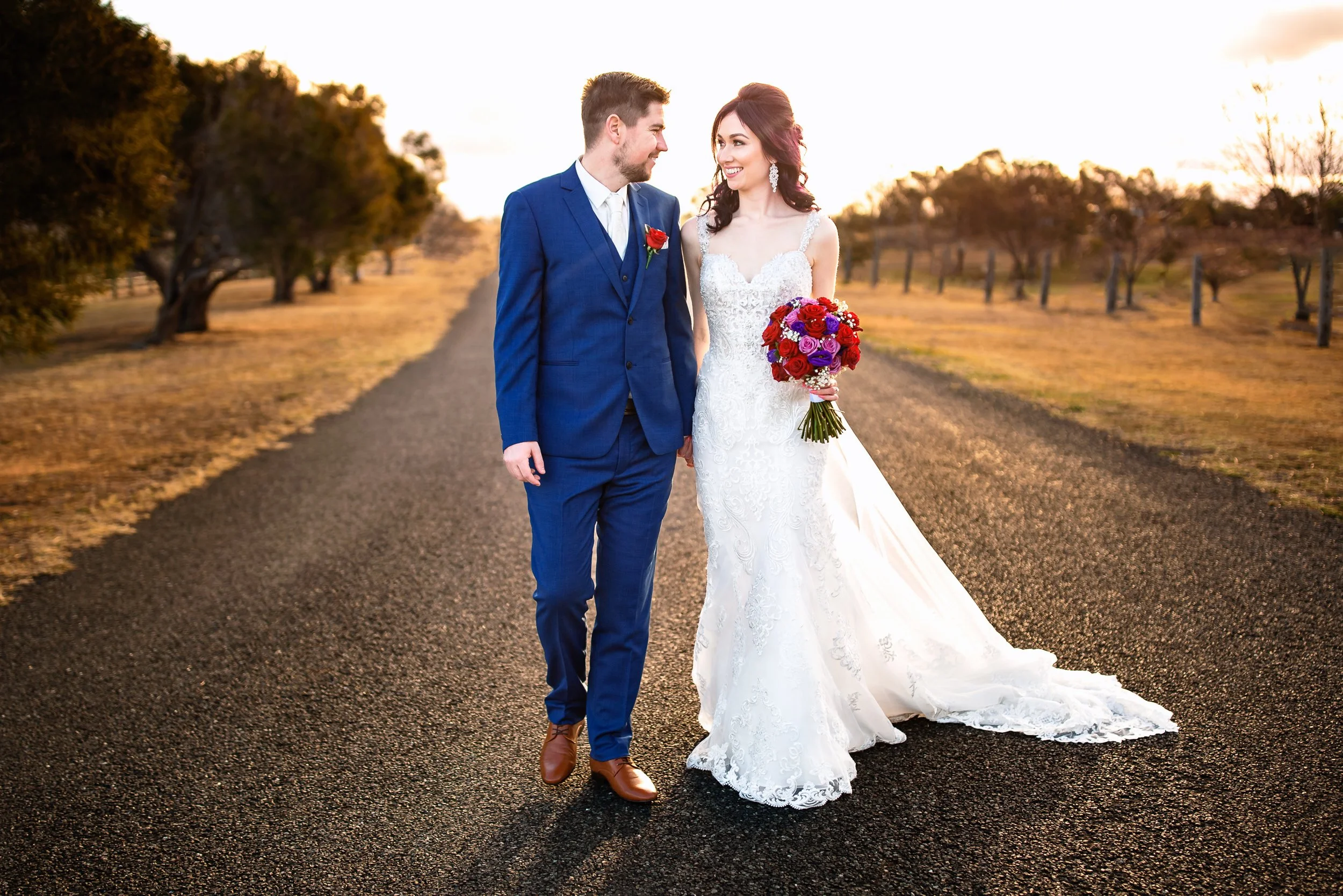 A newlywed couple walking hand in hand in Pokolbin, Hunter Valley during sunset, with the bride in a white lace wedding gown holding a bouquet of red, purple, and white flowers, and the groom in a blue suit with a white shirt and brown shoes.