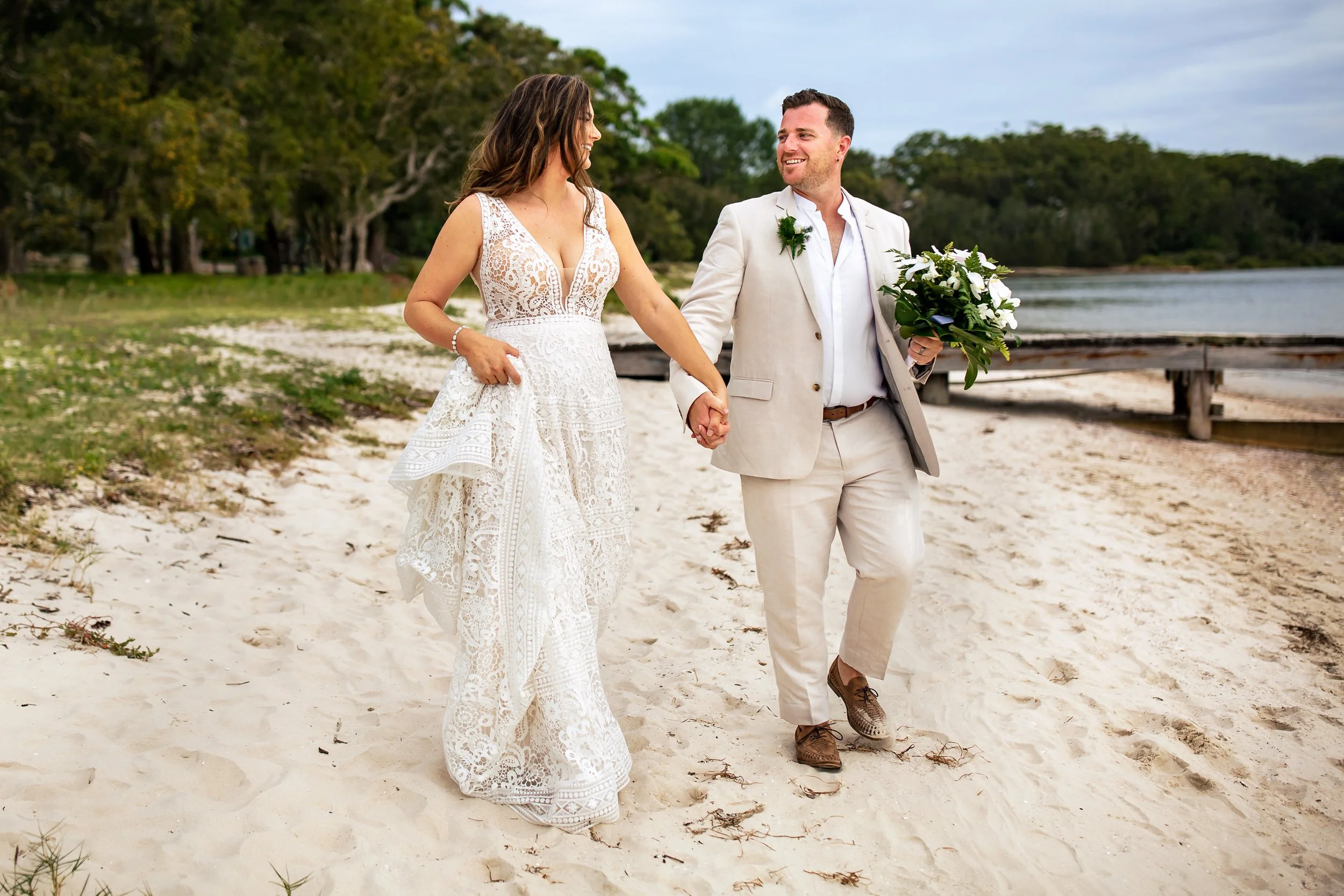 A bride and groom walking along Sunset Beach, Port Stephens, with Pearson Park and the Karuah River in the background.