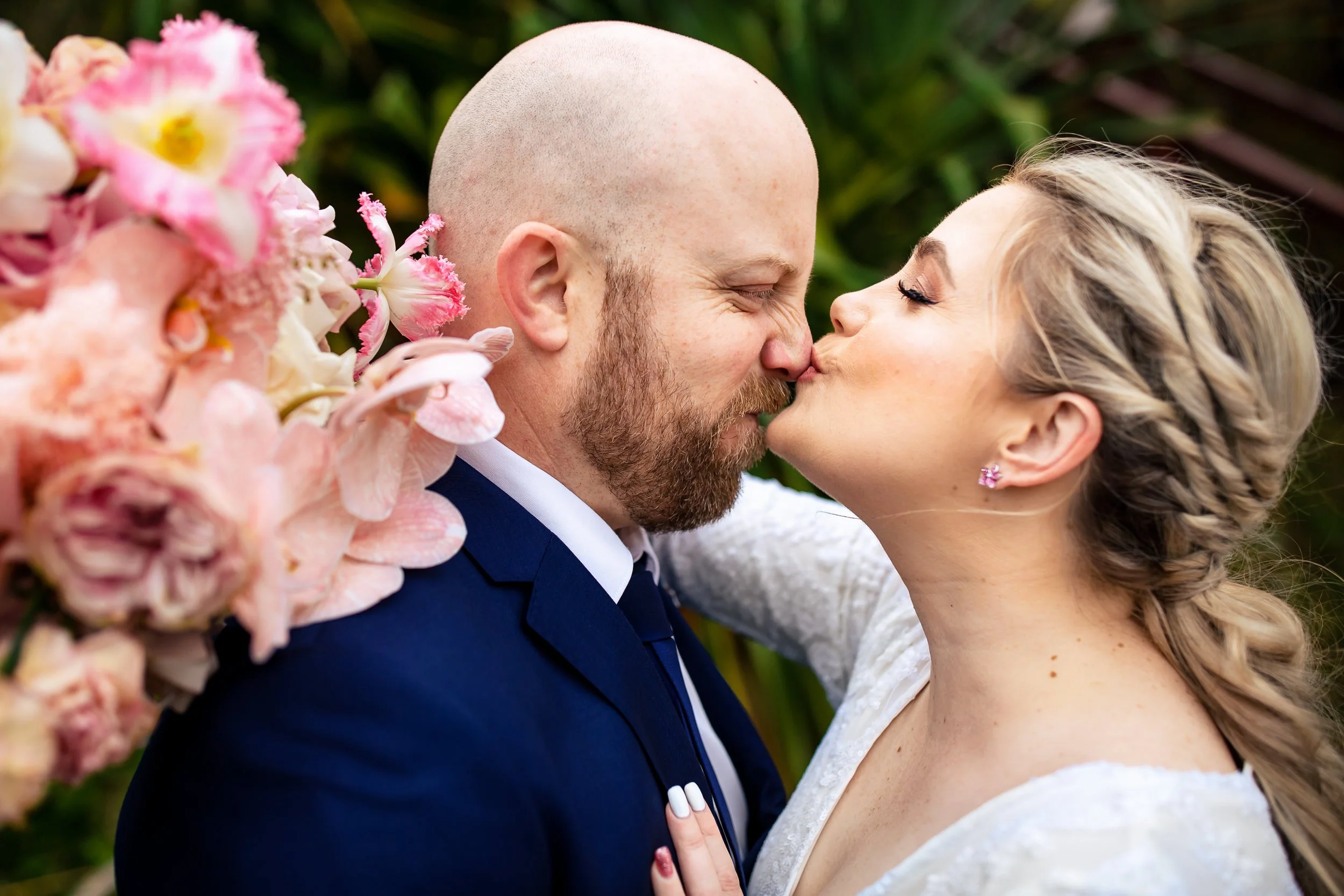 A couple in wedding attire sharing a kiss outdoors, surrounded by pink flowers and greenery. Photos by Port Stephens wedding photographers, Lynn & Justin.