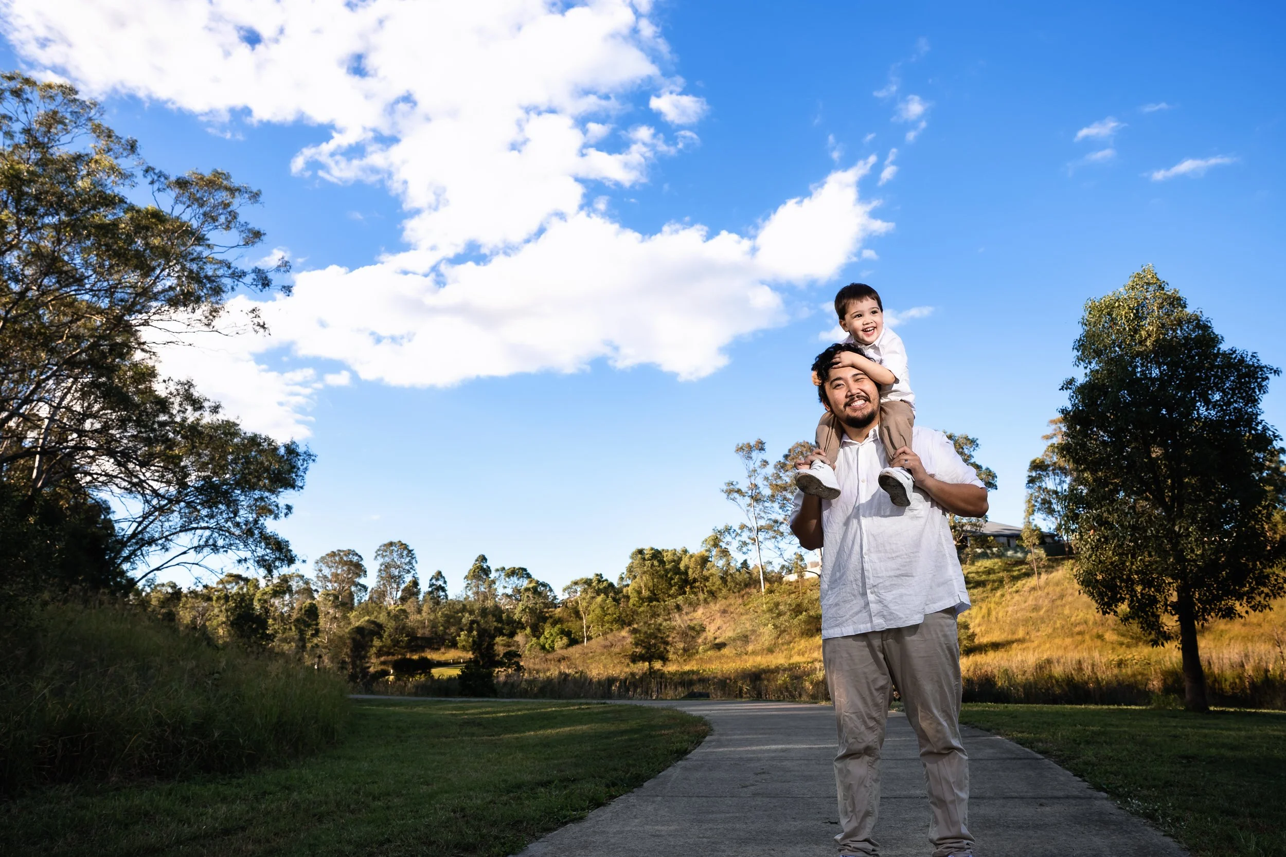 A man carrying a smiling young boy on his shoulders along a park pathway with trees and a blue sky with clouds in the background.