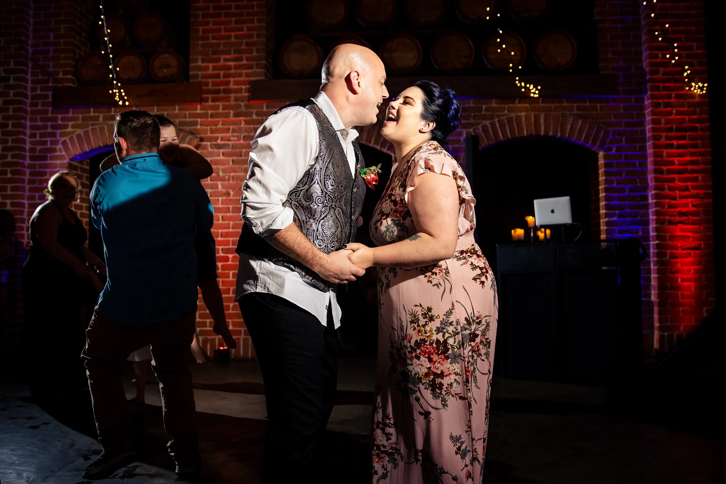 A bride and groom dance together at a wedding reception in a brick venue, with other guests in the background and string lights hanging.